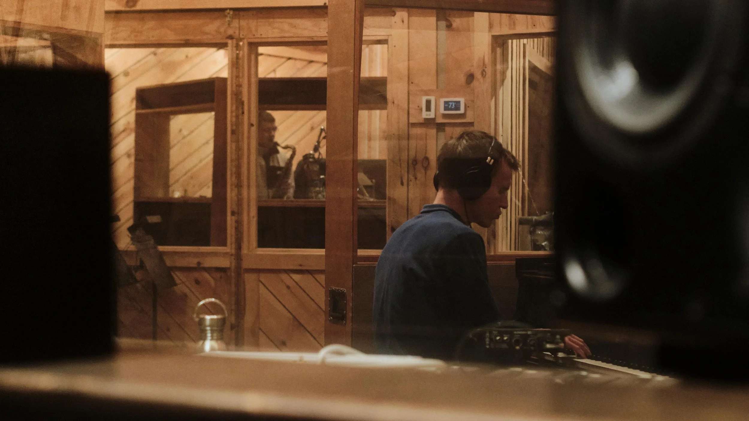 Recording studio interior with a person playing piano and another person in a soundproof room playing saxophone.