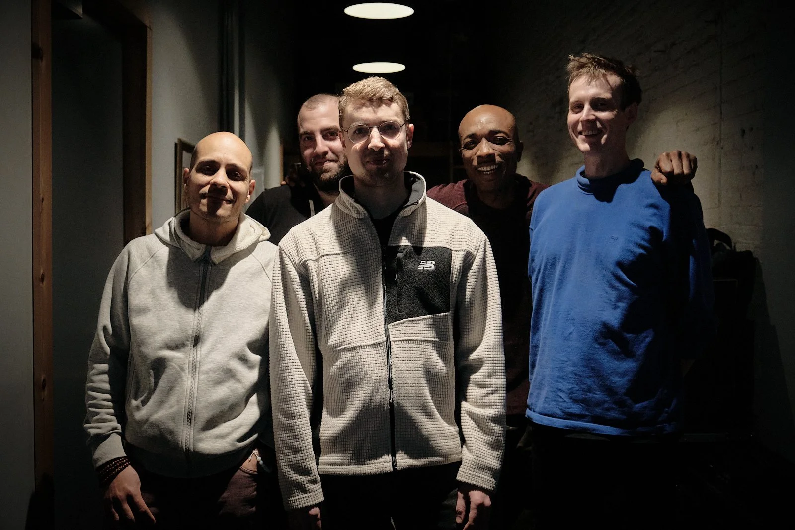 Bassist Chris Tordini, saxophonist Alain Metrailler, pianist Elias Stemeseder, harmonicist Gregoire Maret and drummer Eric McPherson standing together inside the Bunker, smiling and posing for a group photo, with overhead circular lights above them.