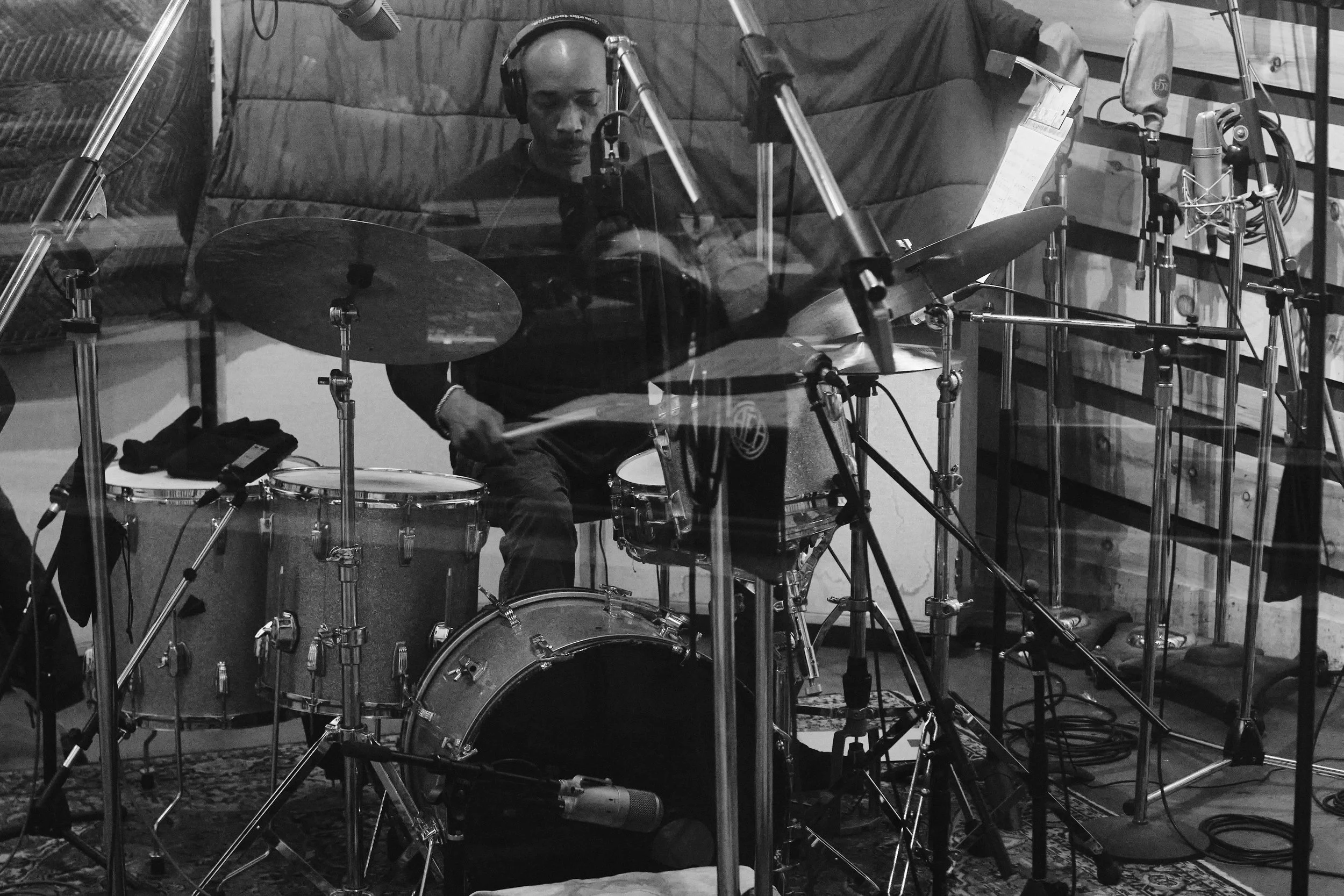 Black and white image of a drummer playing a drum kit in a recording studio with microphones set up around the kit.