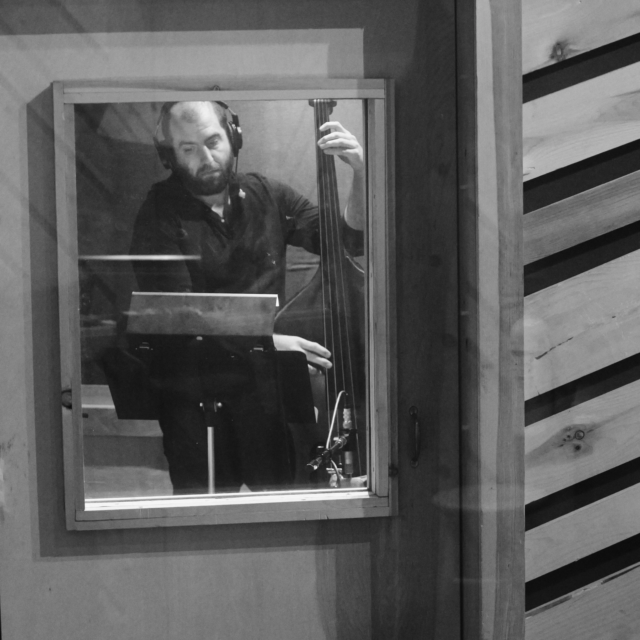 Black and white photo of a musician playing a double bass in a recording studio booth, wearing headphones.