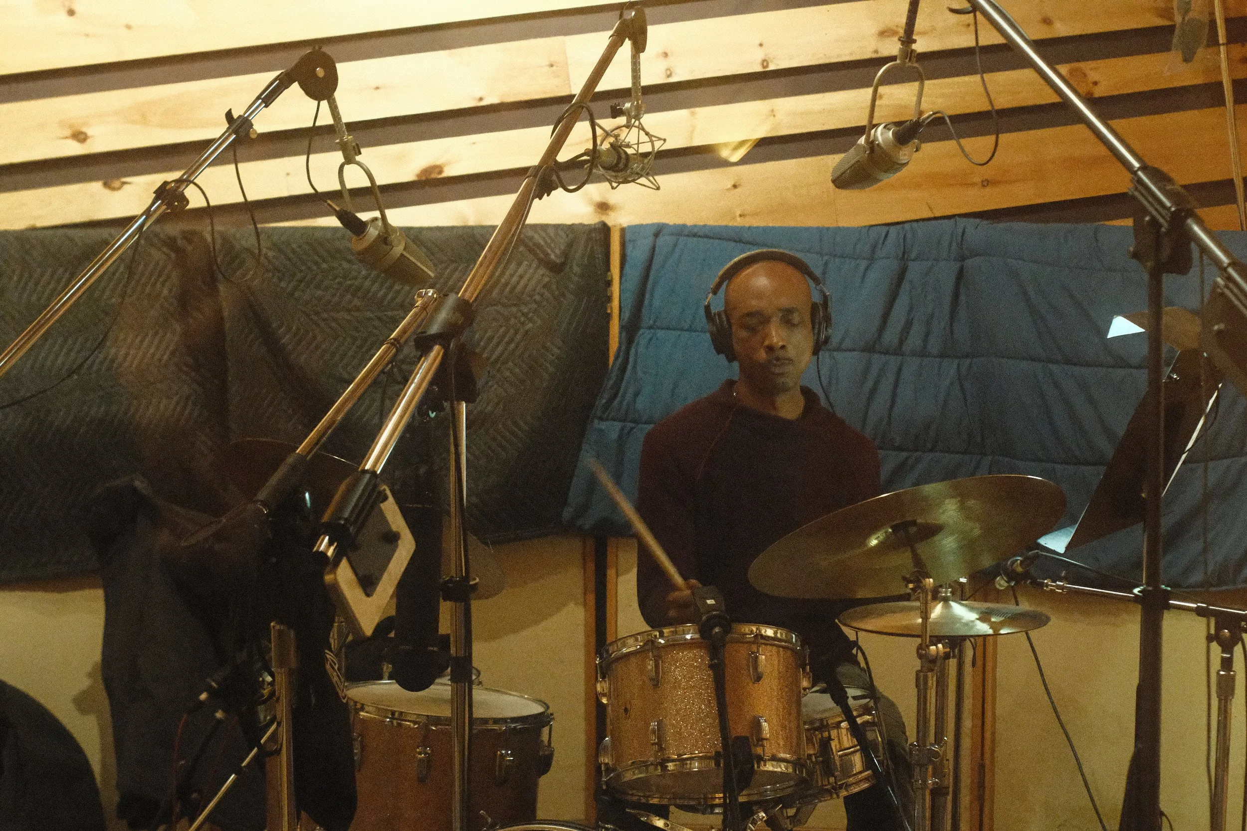Drummer wearing headphones in a recording studio with drum kit and microphones.