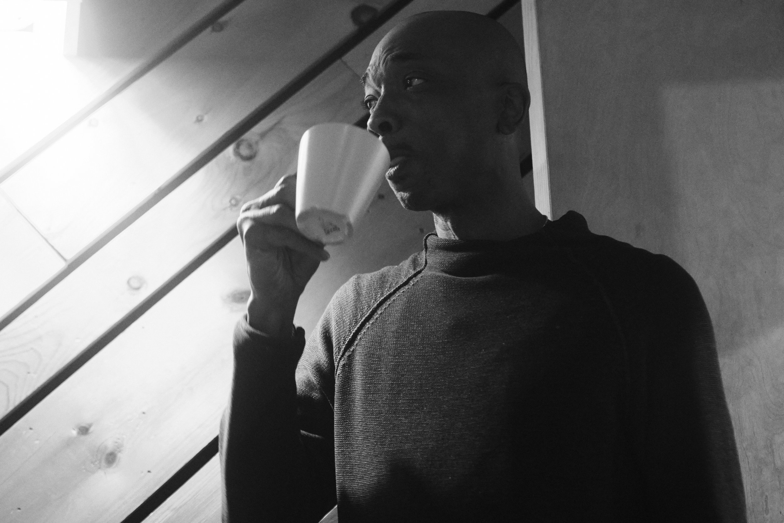 Black and white photo of a man drinking from a mug indoors with wood panel background.