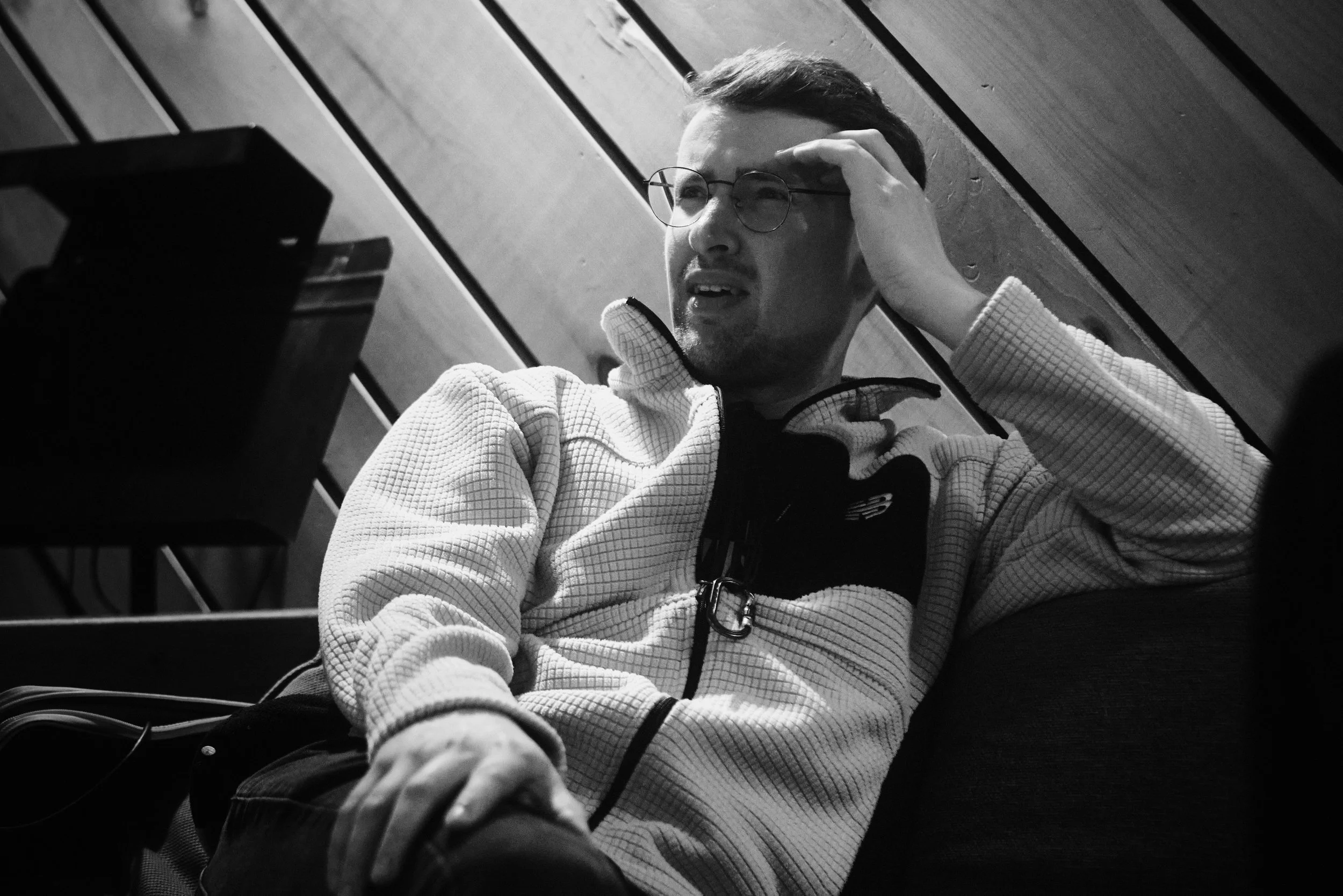 Black and white photo of Alain Metrailler in glasses and a textured jacket, seated on a couch, with slatted wooden wall in background.