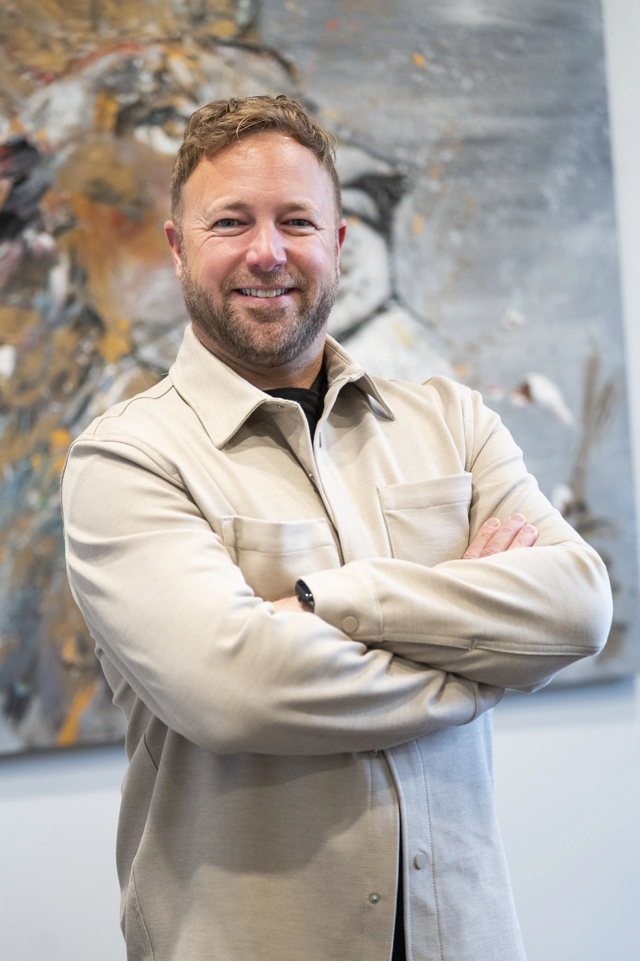 Homme d'affaires souriant portant un costume bleu foncé et une chemise blanche à petits motifs, debout à côté d'un escalier intérieur avec large fenêtre en arrière-plan.