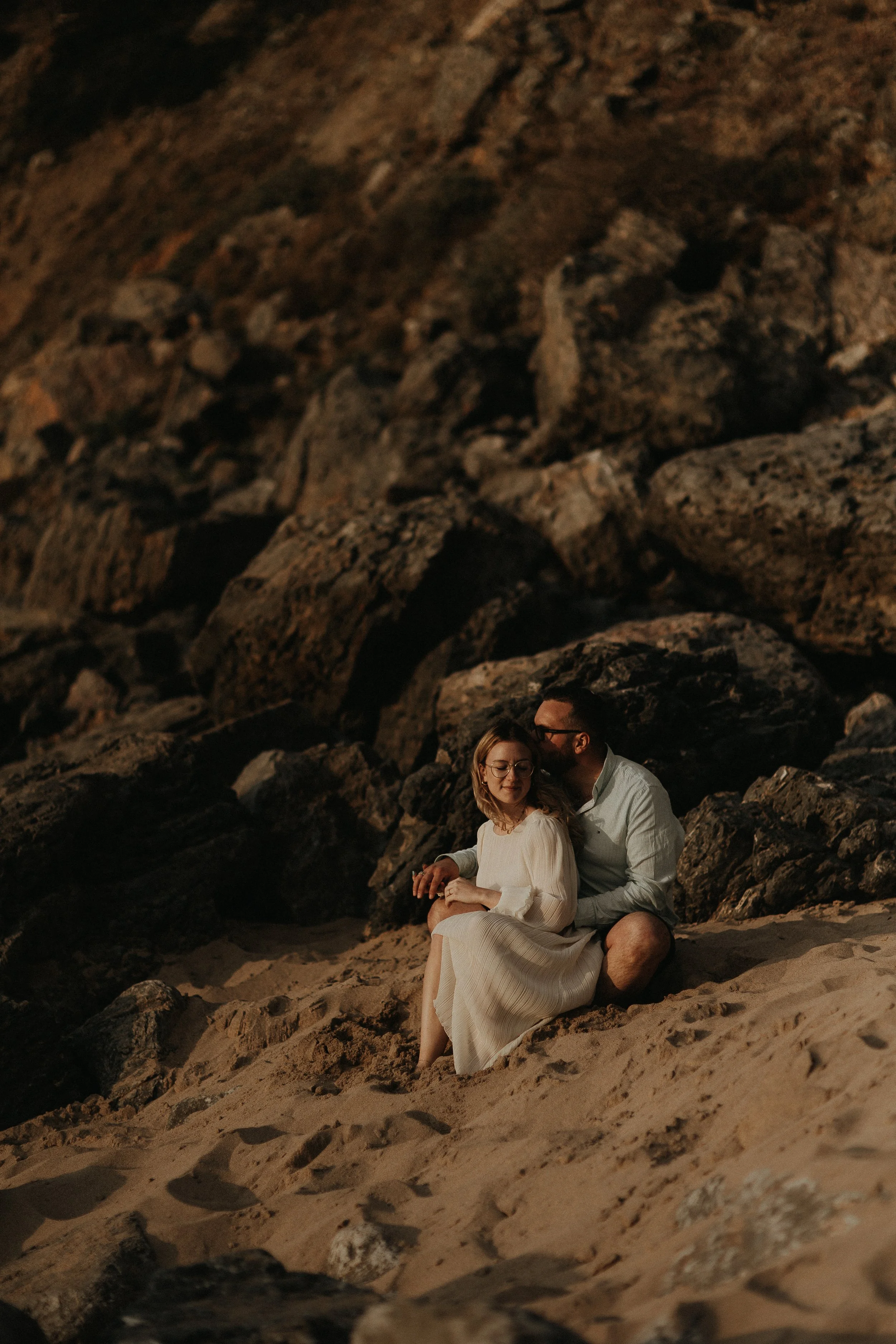 A couple sitting on a sandy beach surrounded by large rocks.