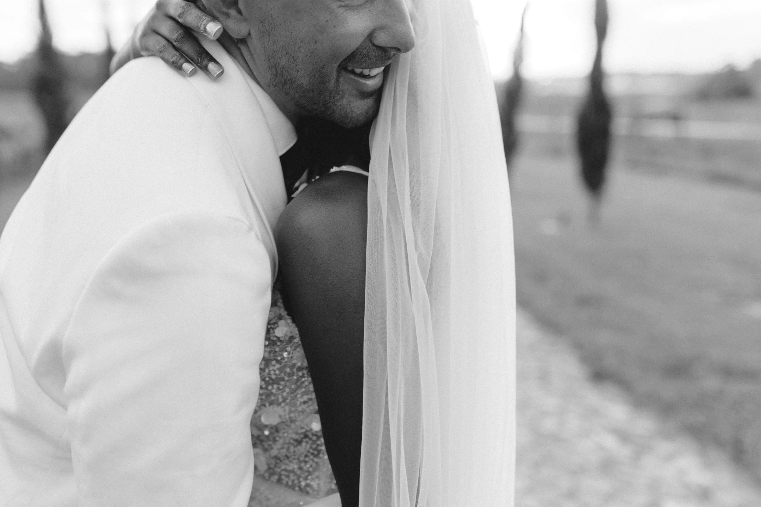 A close-up black and white photo of a couple hugging, with the woman wearing a veil and a dress with embellishments, and the man in a white suit. The background is blurred. The moment appears tender and joyful.