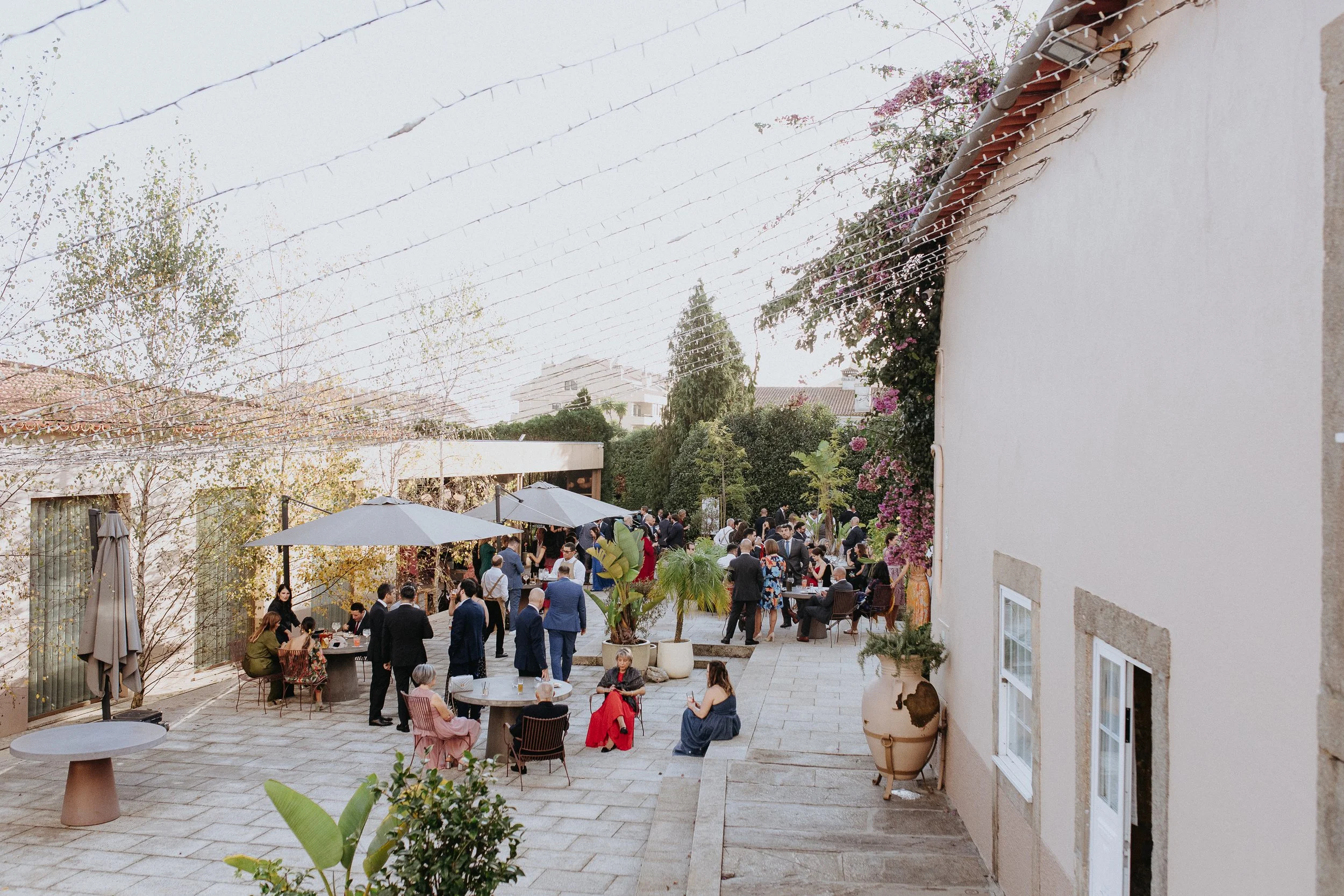 People gathered in an outdoor courtyard for a social event, some standing and talking, others sitting at tables with umbrellas, surrounded by potted plants and flowering bushes.