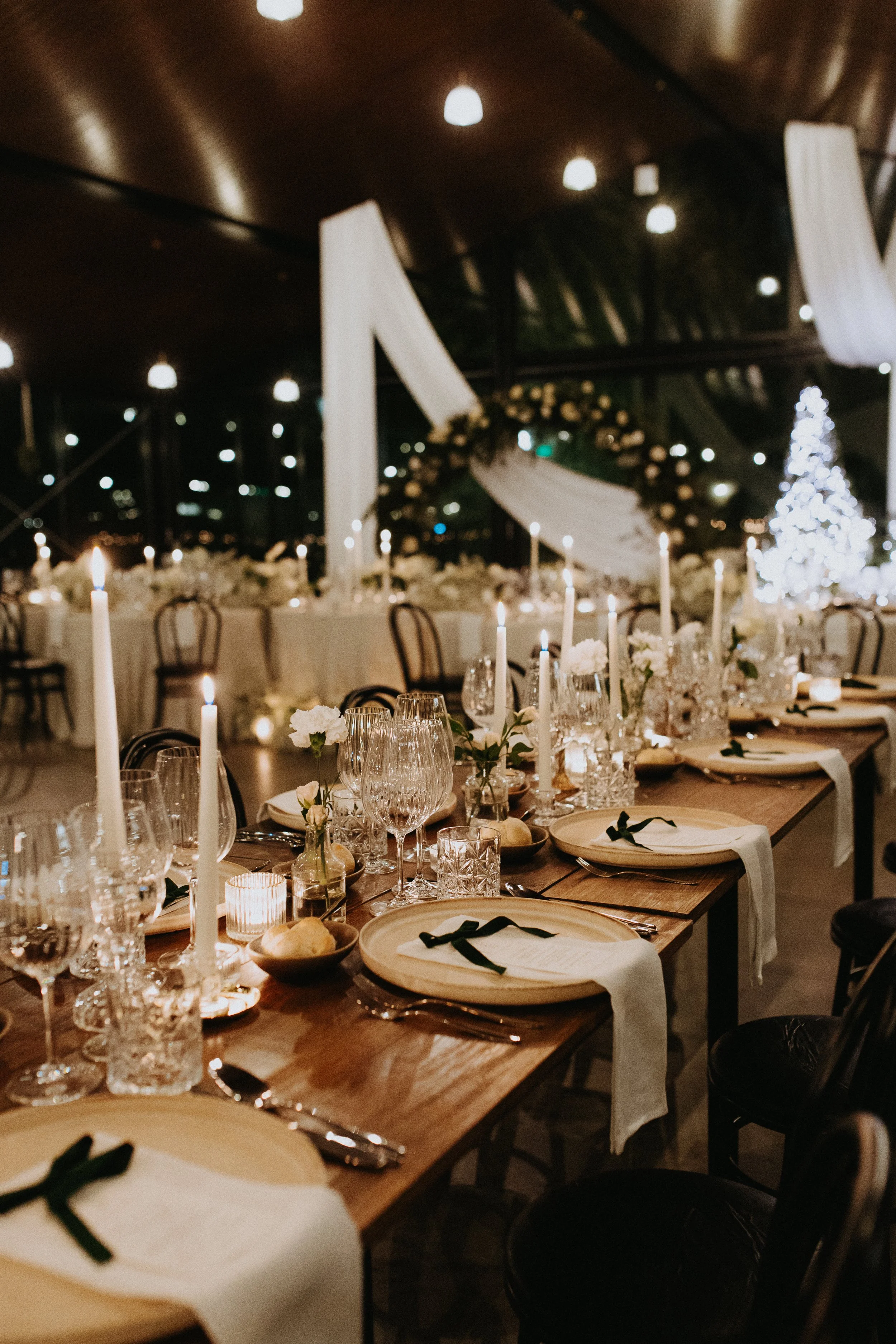 Mesa de jantar decorada com velas, pratos, talheres, copos e guardanapos em um ambiente com iluminação suave, no estilo de festa de fim de ano com árvore de Natal iluminada ao fundo.