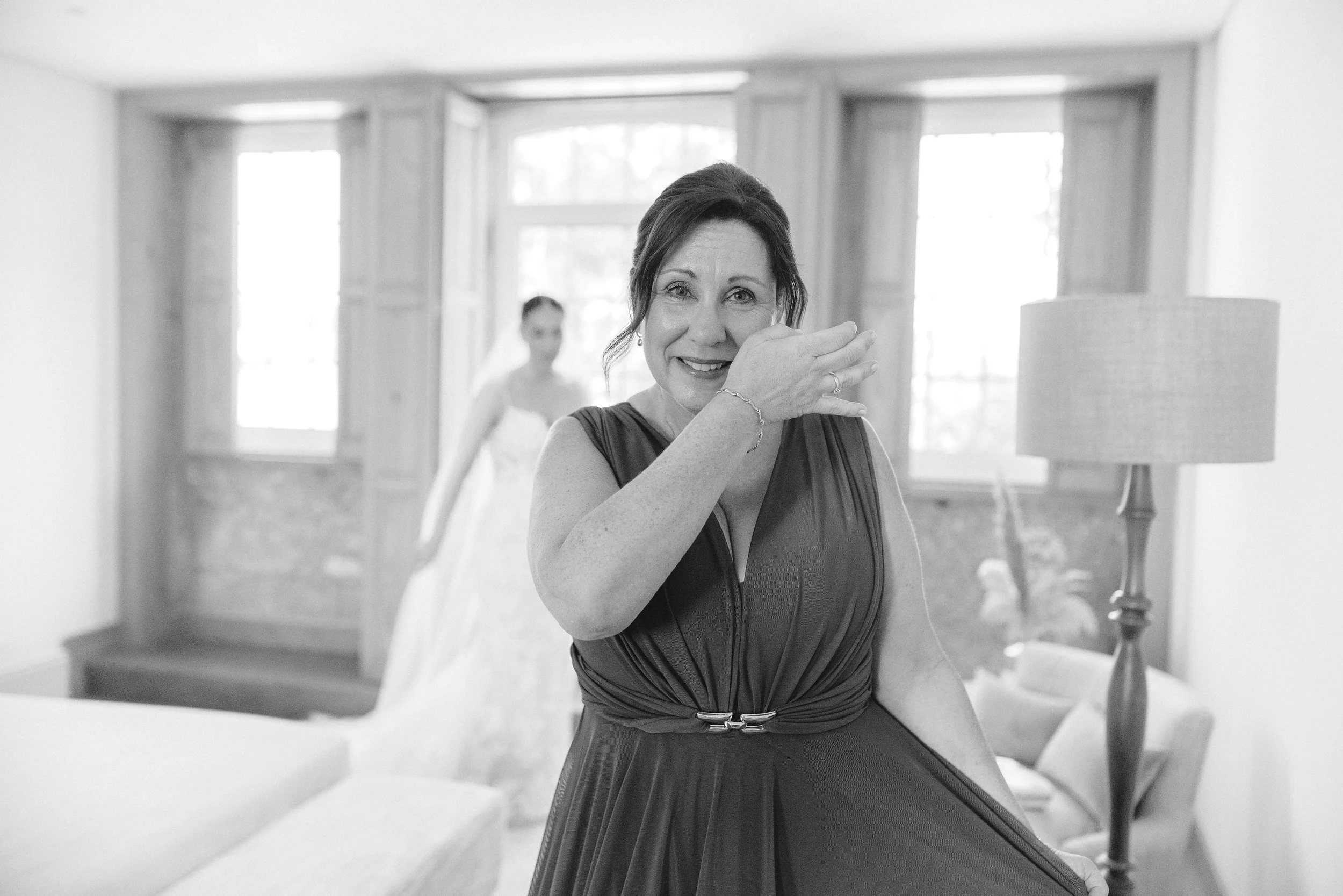 A woman in an elegant dress smiling and wiping tears from her eye in a well-lit room with a bride in a wedding dress in the background.