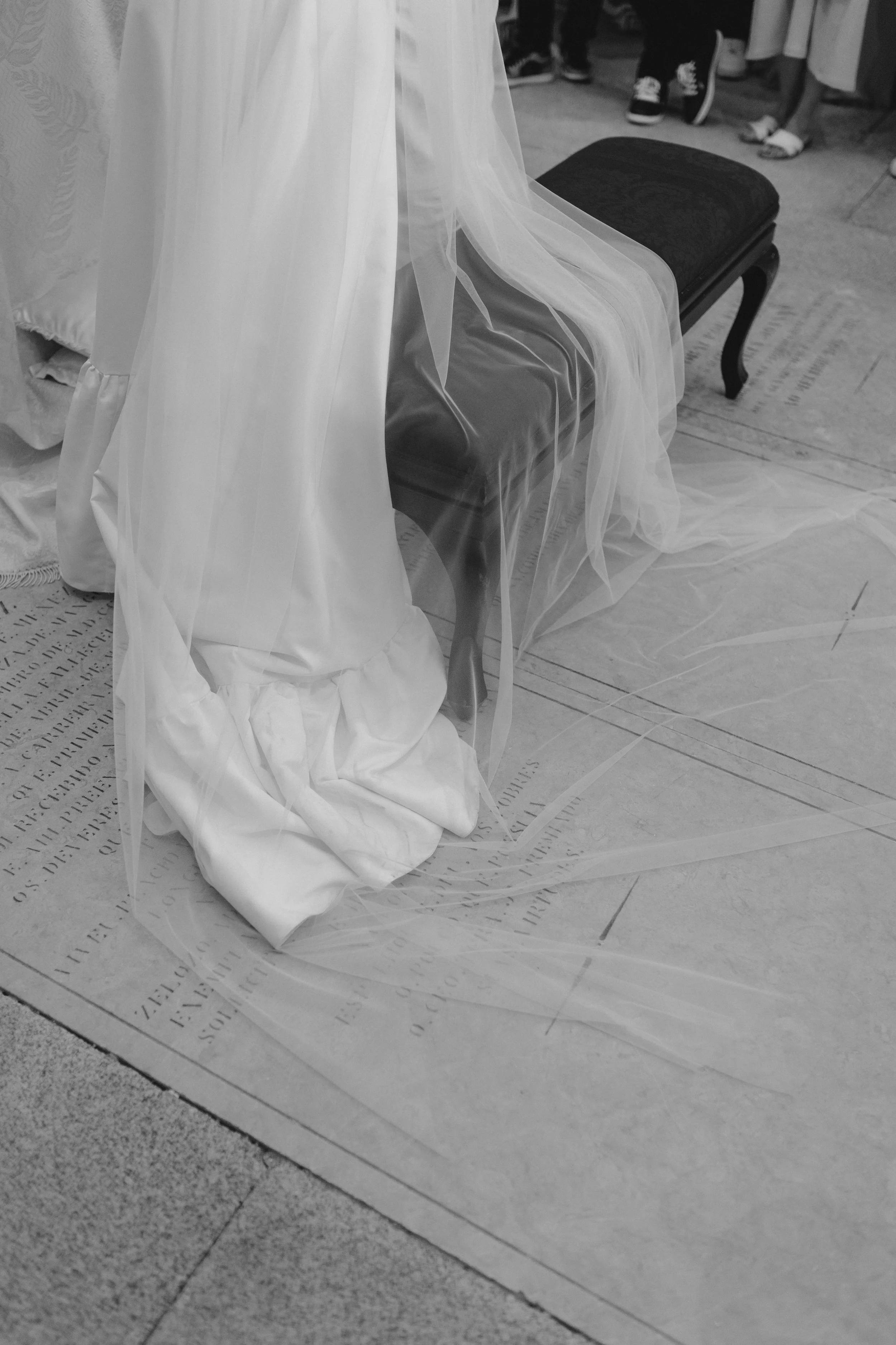 Imagem em preto e branco de uma cadeira com uma peça de roupa branca e um véu, possivelmente em um casamento, com pessoas ao fundo e texto no chão.