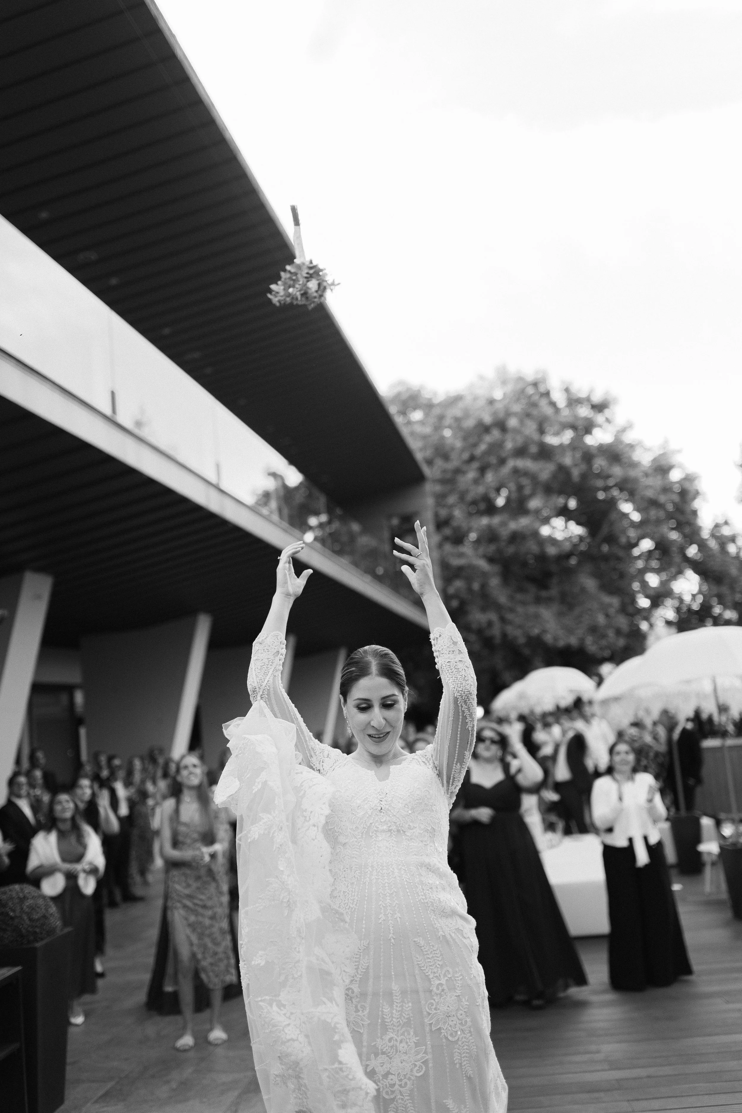 A woman in a lace wedding dress throws her bouquet to a group of women holding umbrellas at an outdoor wedding reception.