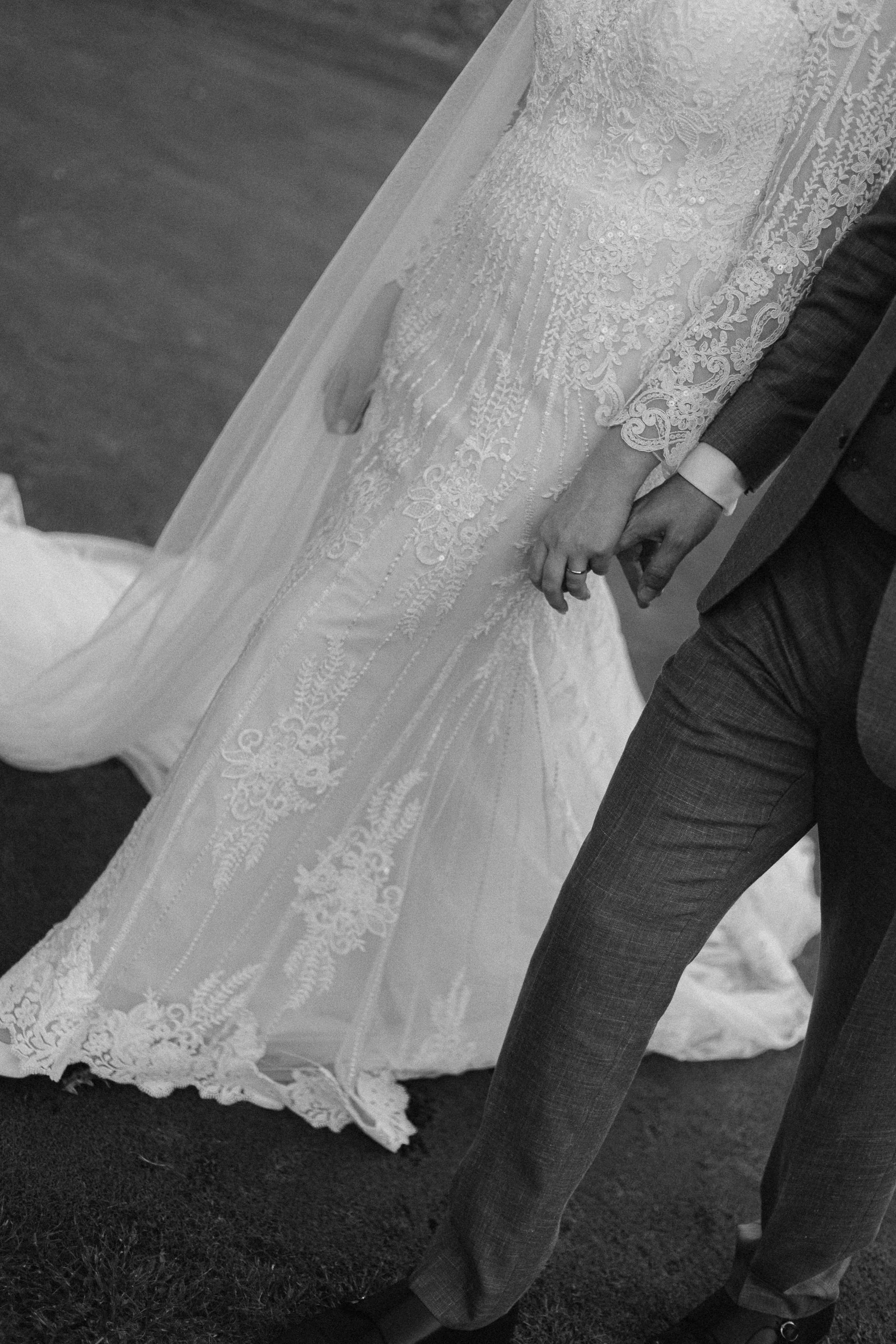 Close-up of a bride and groom holding hands during their wedding ceremony, showing the bride's lace wedding dress and the groom's suit.