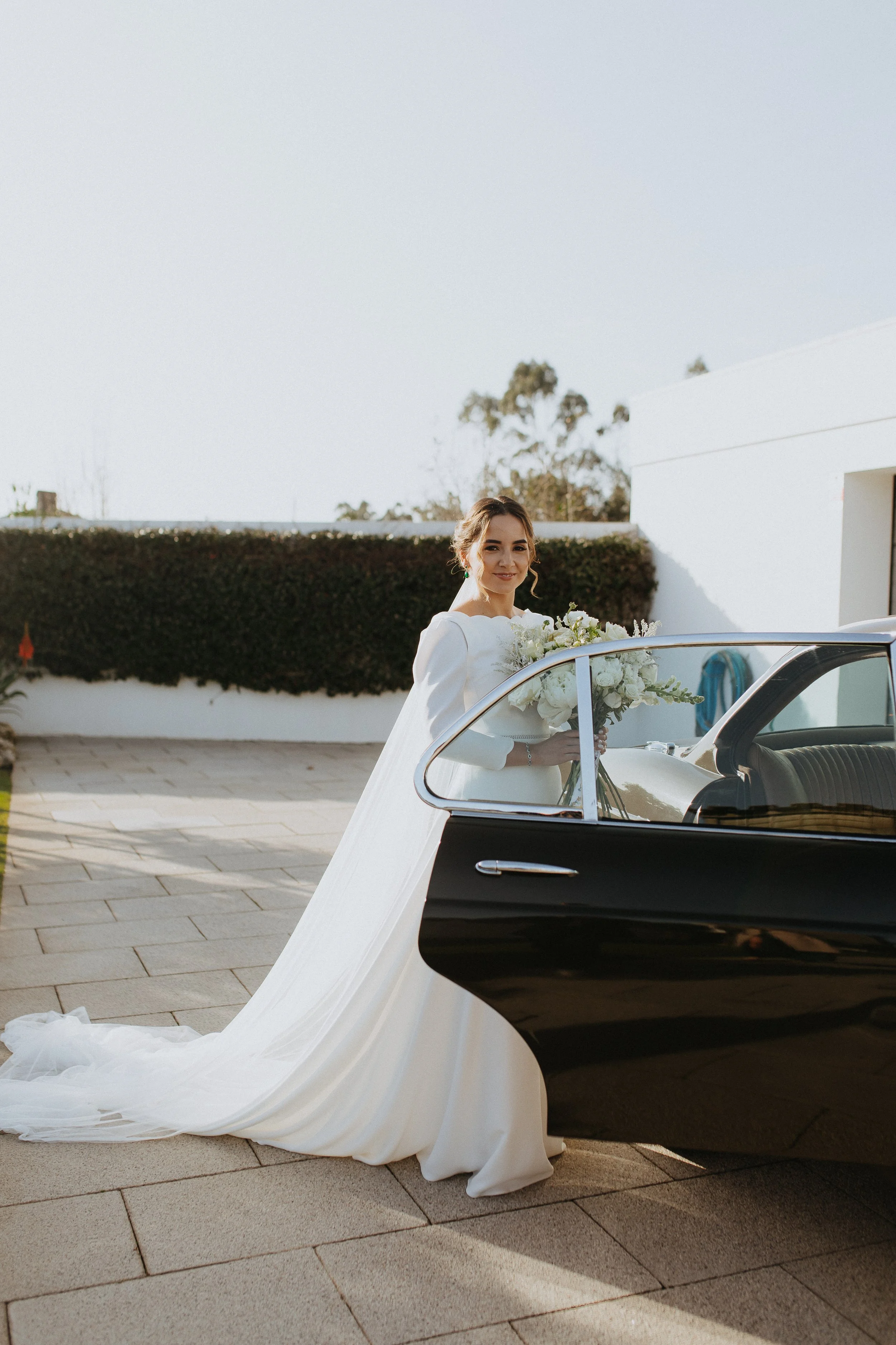 Noivo sorridente com vestido de noiva branco segurando buquê de flores, ao lado de carro clássico preto e branco em ambiente ao ar livre.
