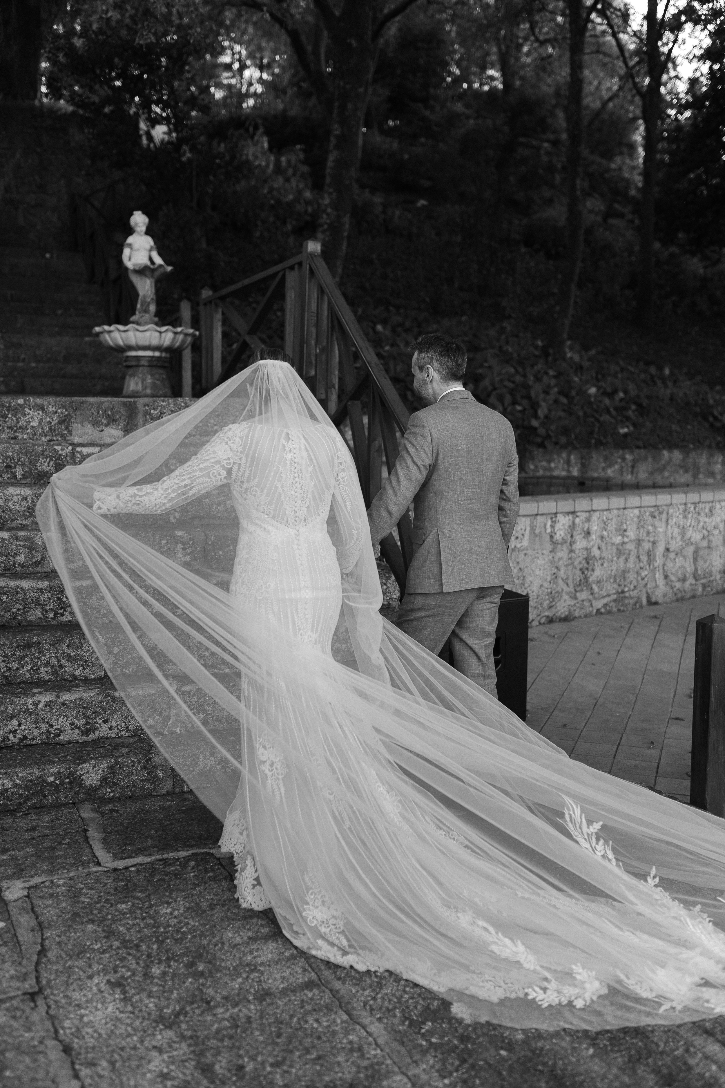 A bride and groom walking up stone stairs outdoors, with the bride wearing a lace wedding dress and a long veil, and the groom in a suit, during a wedding ceremony.