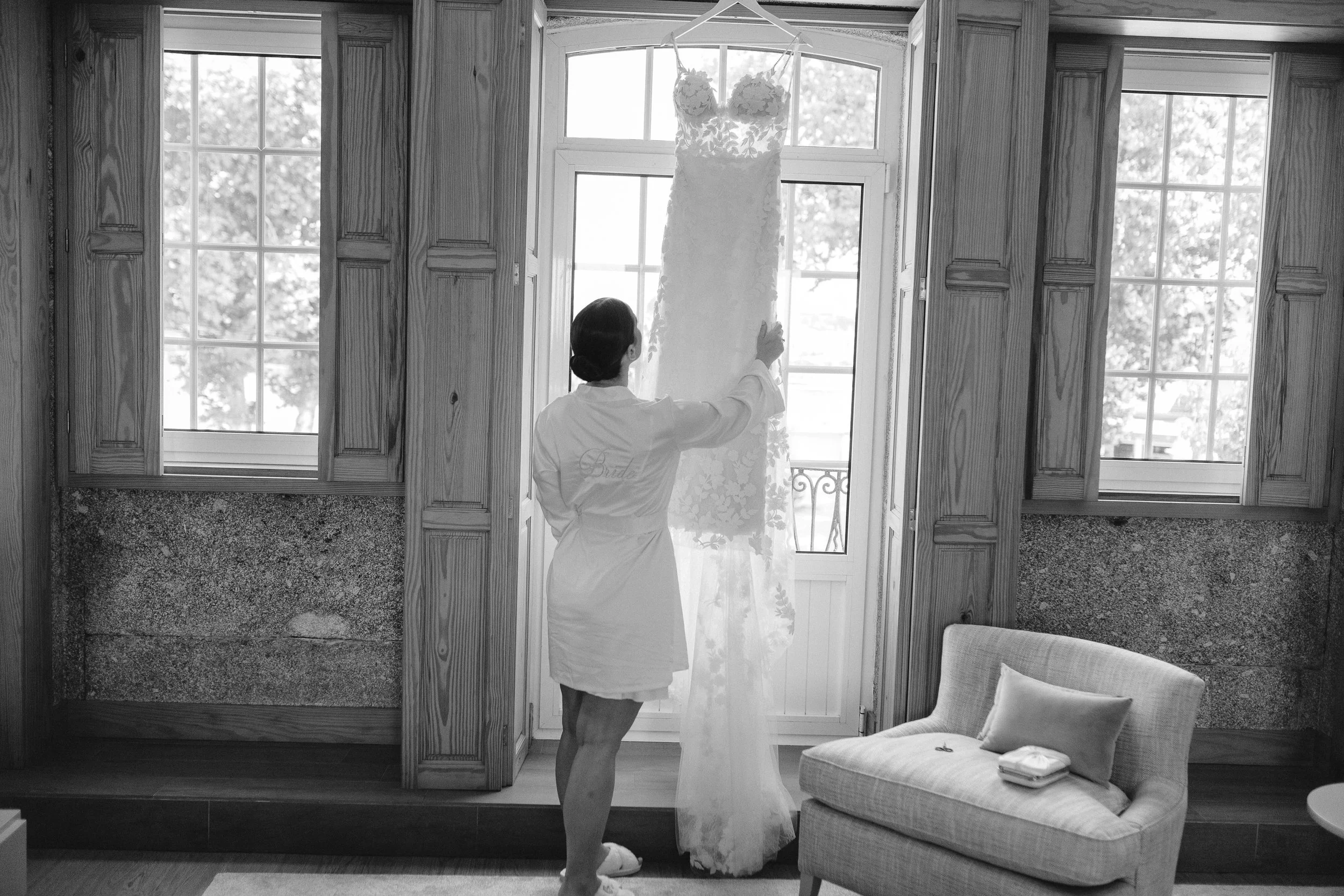 A woman in a robe standing and looking at a wedding dress hanging on a window in a room with wooden paneling and two large windows.