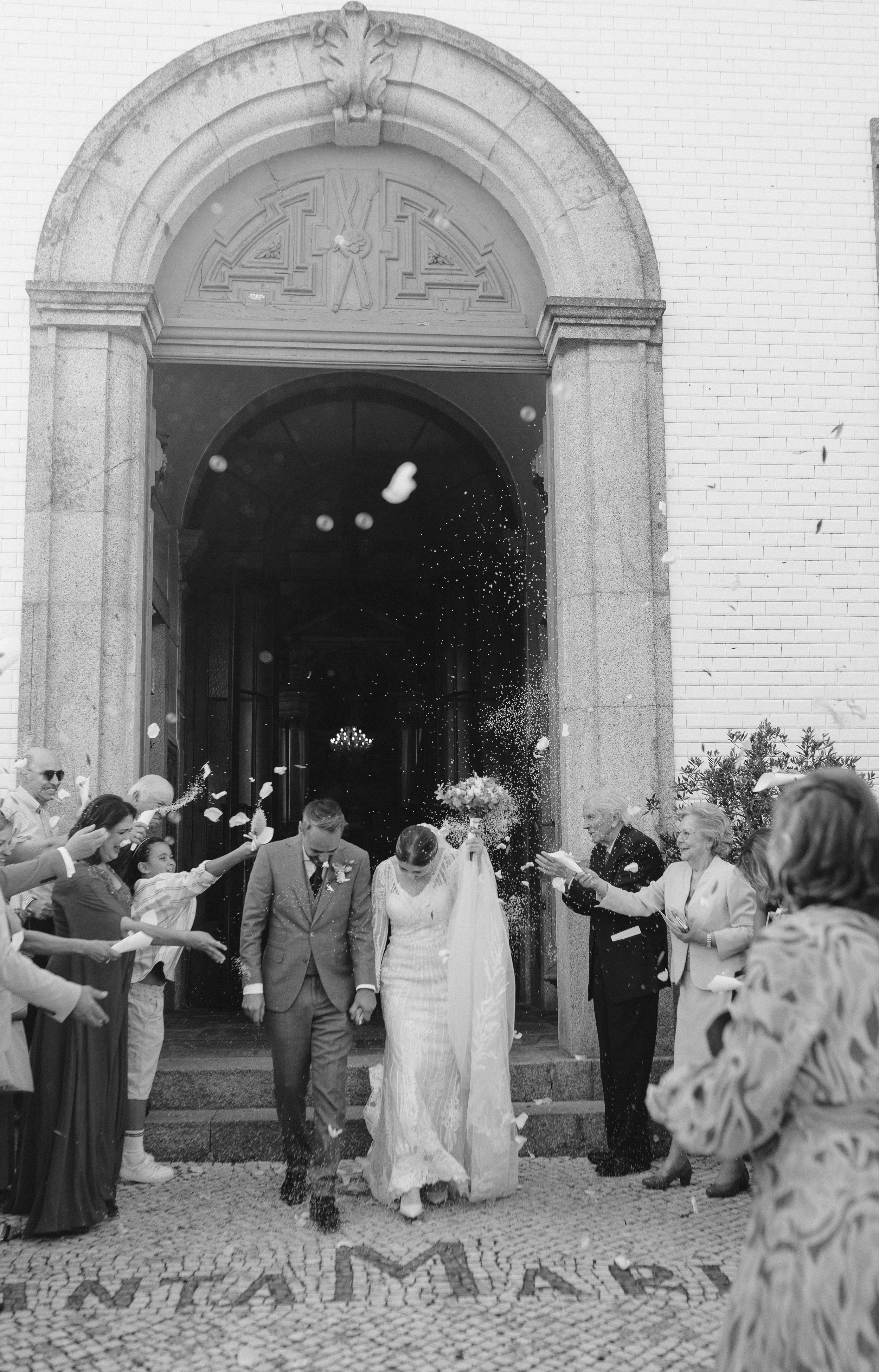 A wedding celebration with a bride and groom leaving a church, guests throwing confetti, and smiling witnesses on a cobblestone street.