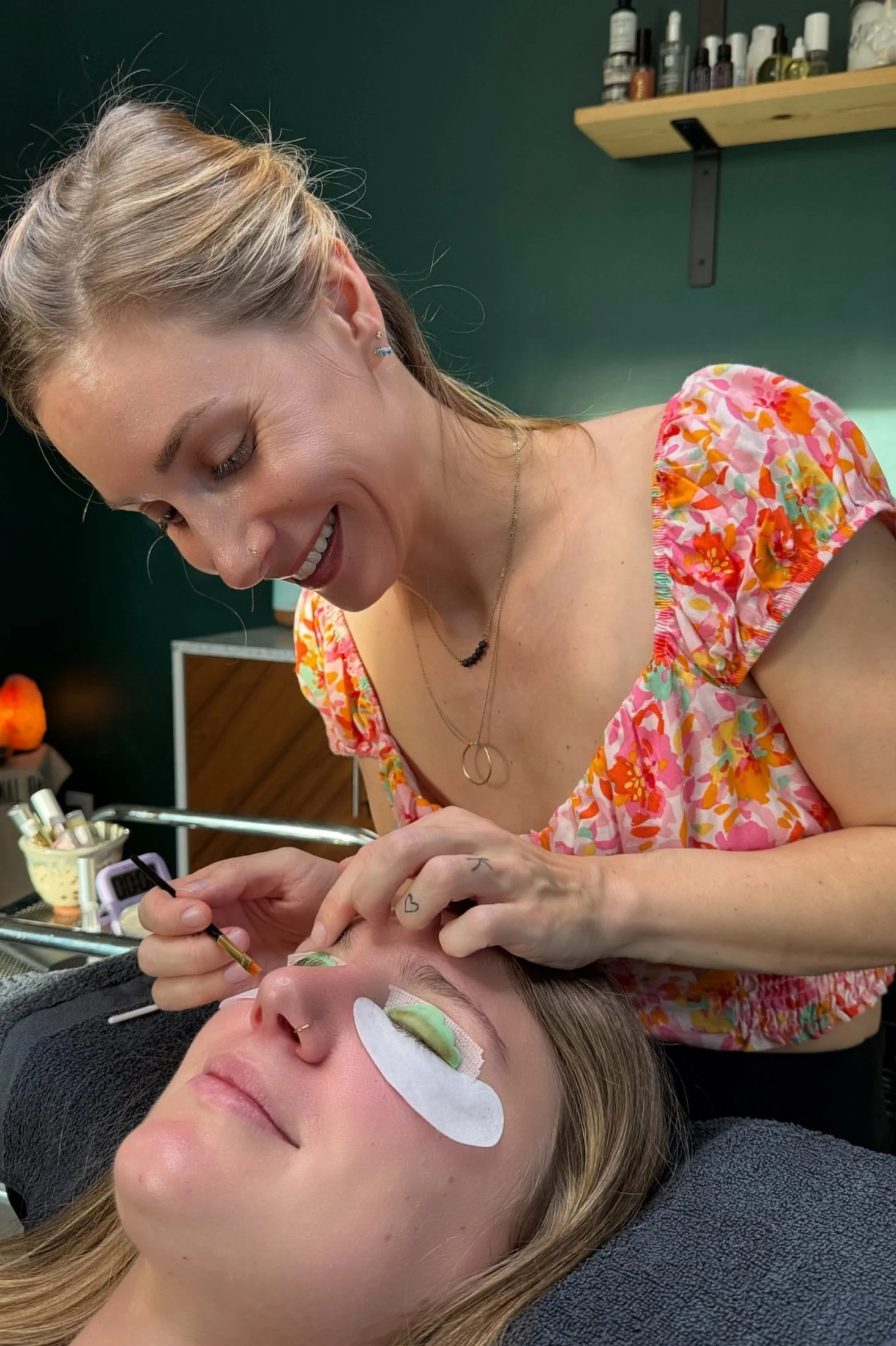 A woman with floral dress receiving an eyelash eyelid extension or lash lift from an aesthetician inside a beauty salon.