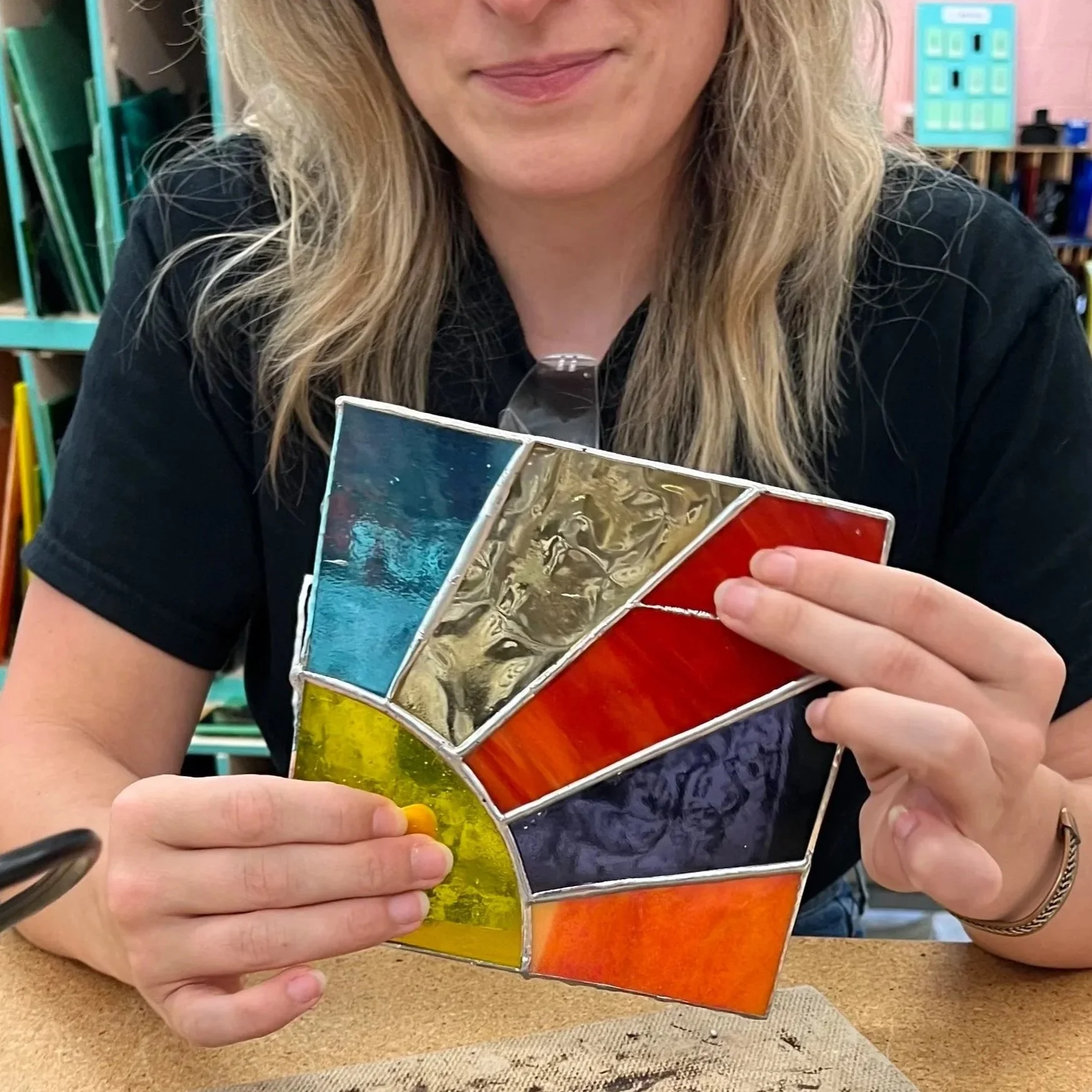 A woman holding a colorful stained glass piece with various rectangular sections in blue, gold, red, green, and orange. The woman is wearing a black shirt and sits at a table in ATL Glassworks with glass shelves in the background.
