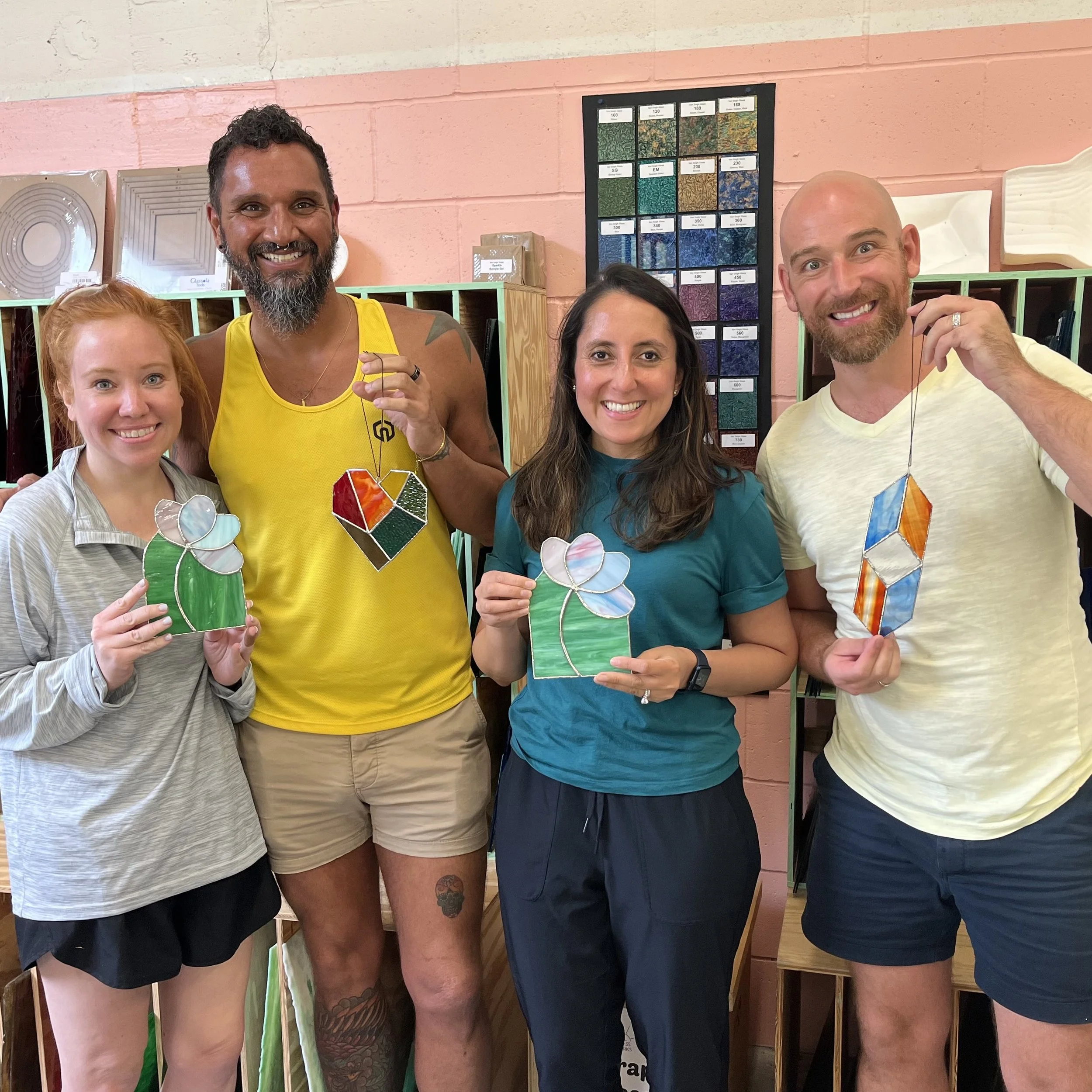 Four people standing indoors, holding colorful stained glass art pieces that they made in an ATL Glassworks Intro to Stained Glass class, smiling at the camera.