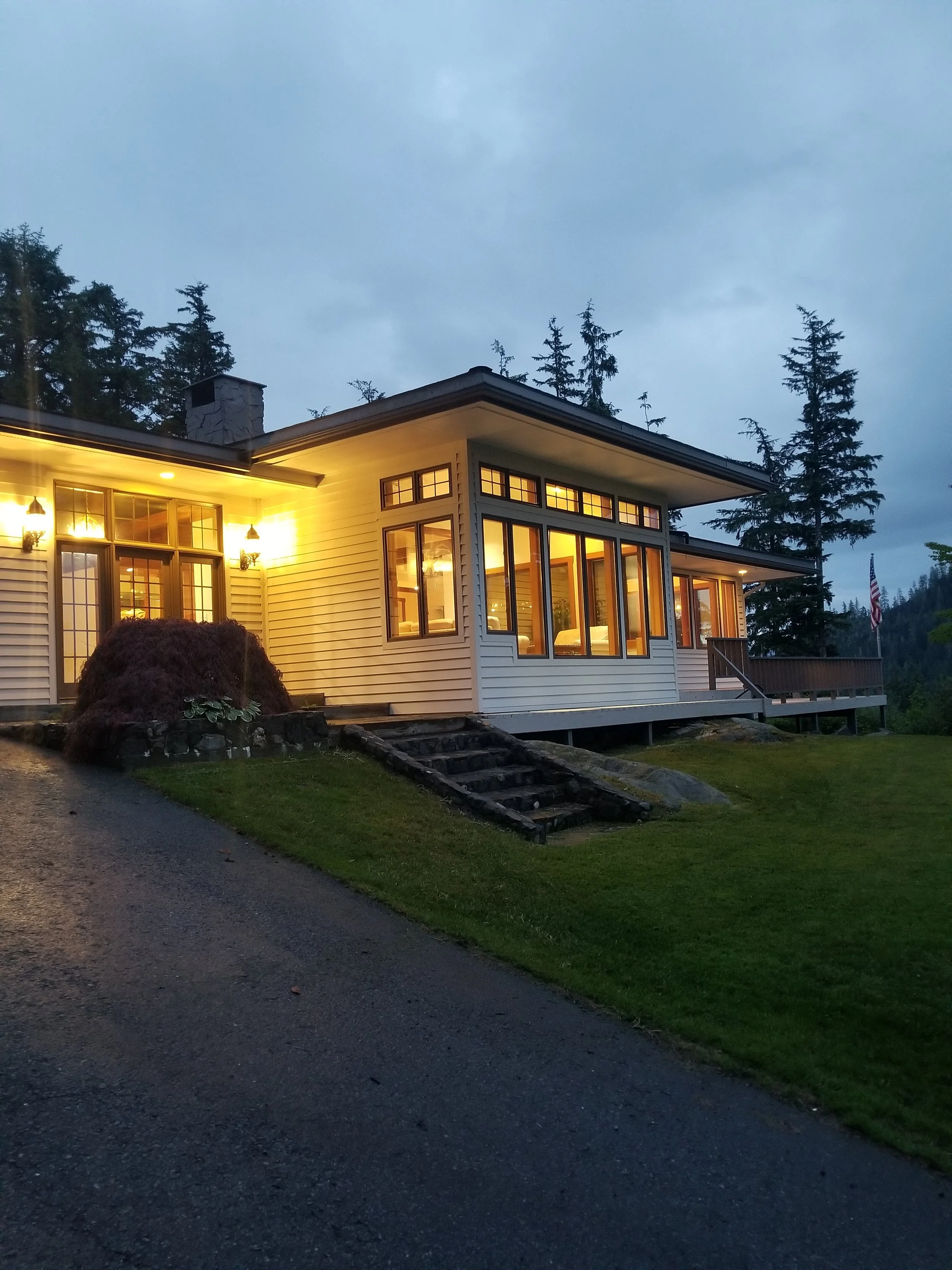 Chrome Chasers fishing lodge exterior at dusk, Wrangell Alaska, with warm interior light glowing through floor-to-ceiling windows and Sitka spruce forest in the background