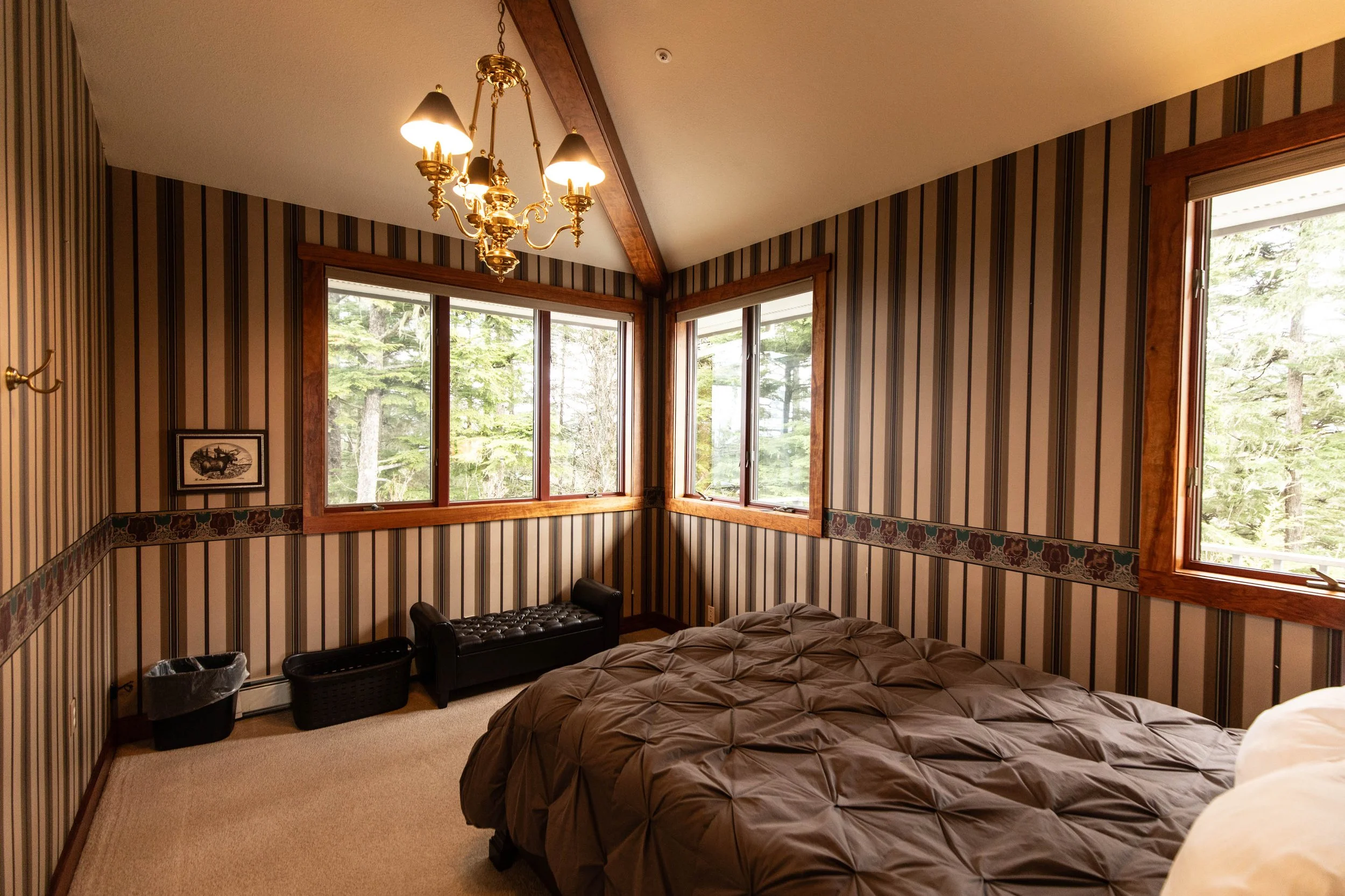 Guest bedroom at Chrome Chasers lodge in Wrangell Alaska featuring vaulted beamed ceiling, brass chandelier, and Brazilian tigerwood window trim with Sitka spruce forest views