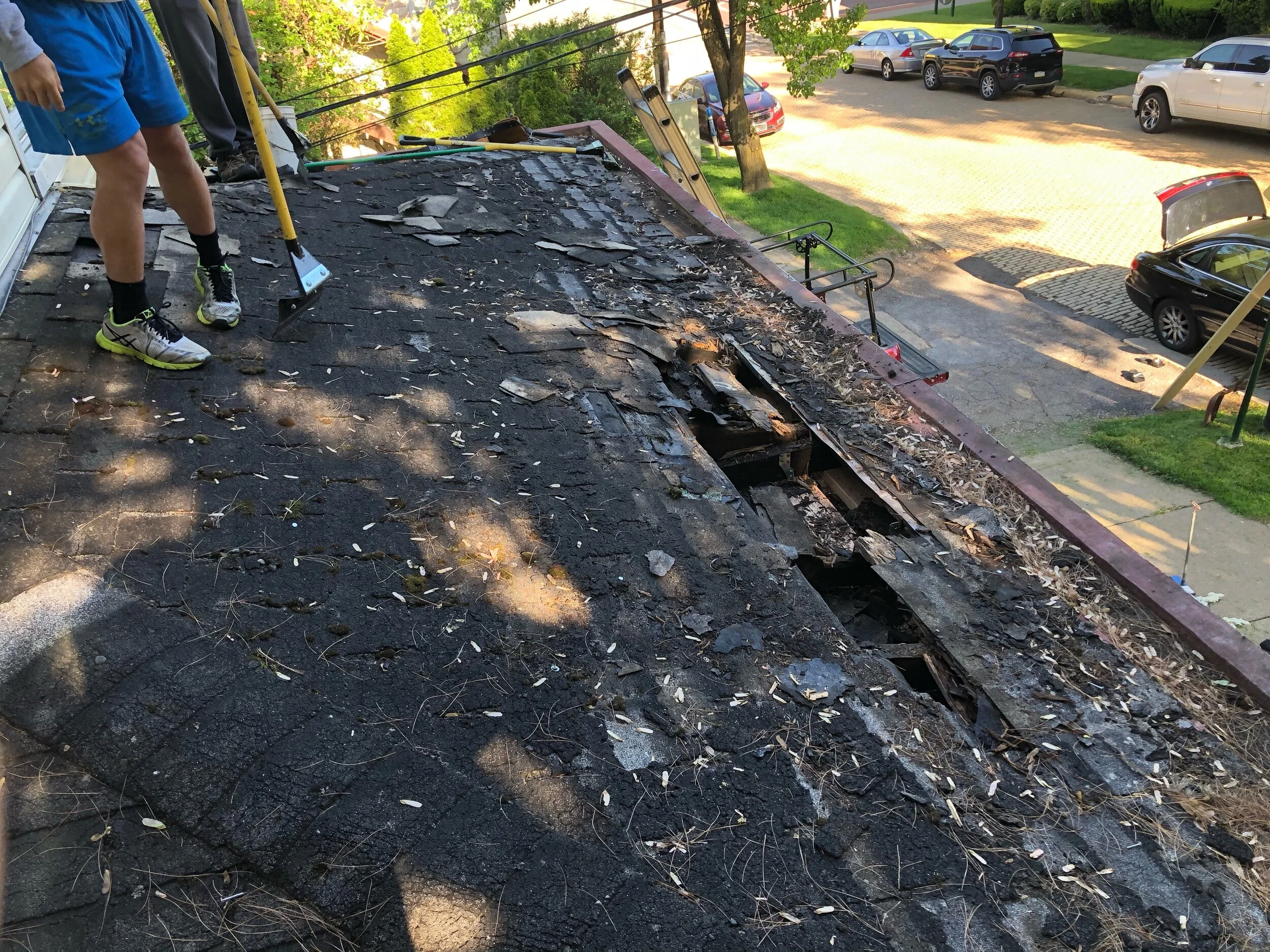 View of a damaged rooftop with missing shingles, debris, and tools, overlooking a parking lot filled with cars and a tree in the background, in a suburban neighborhood during daylight.