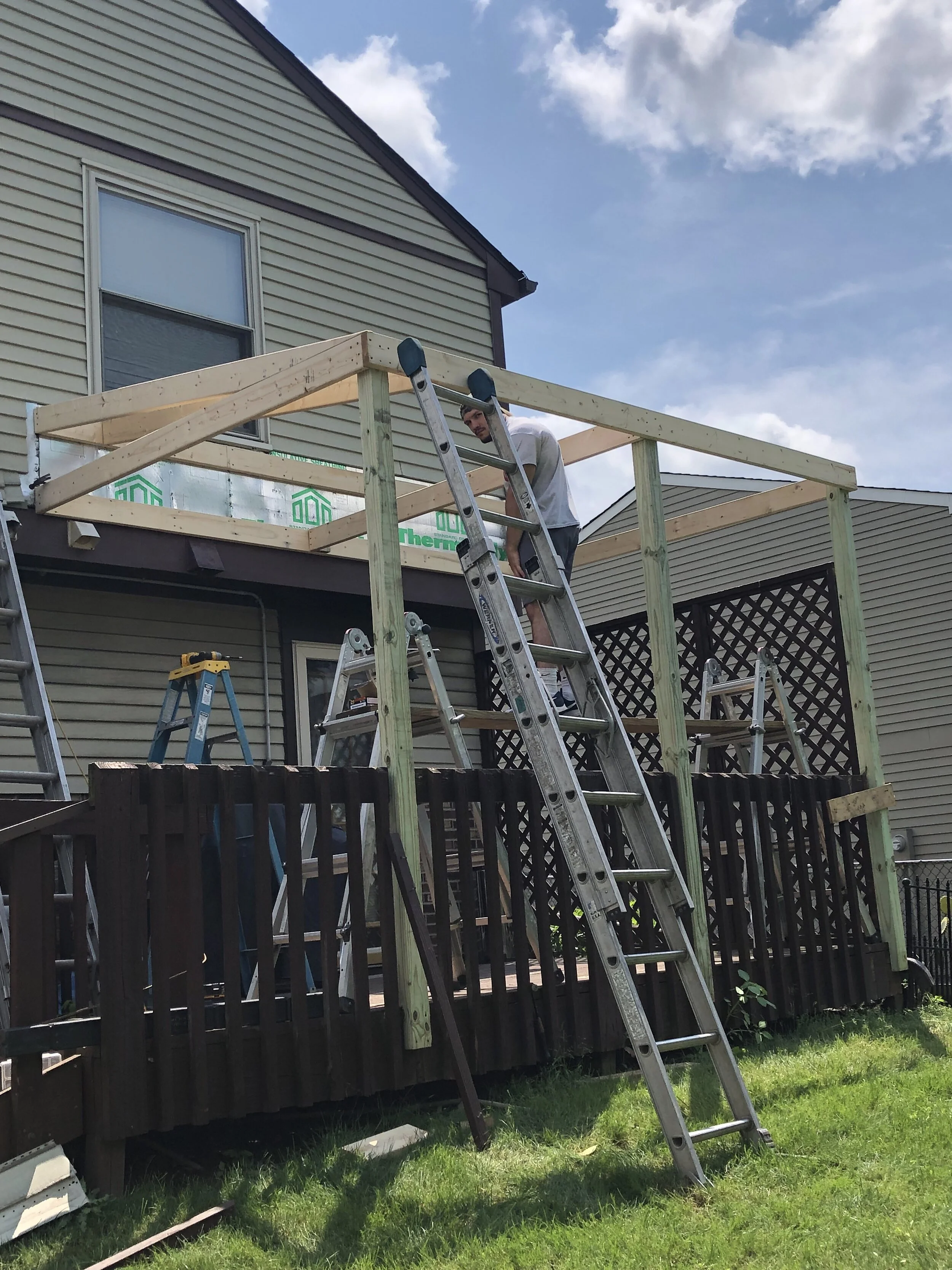 A man working on a wooden deck under construction, using a ladder, with a two-story house with siding and a window in the background, green grass in the foreground, and a partly cloudy sky.