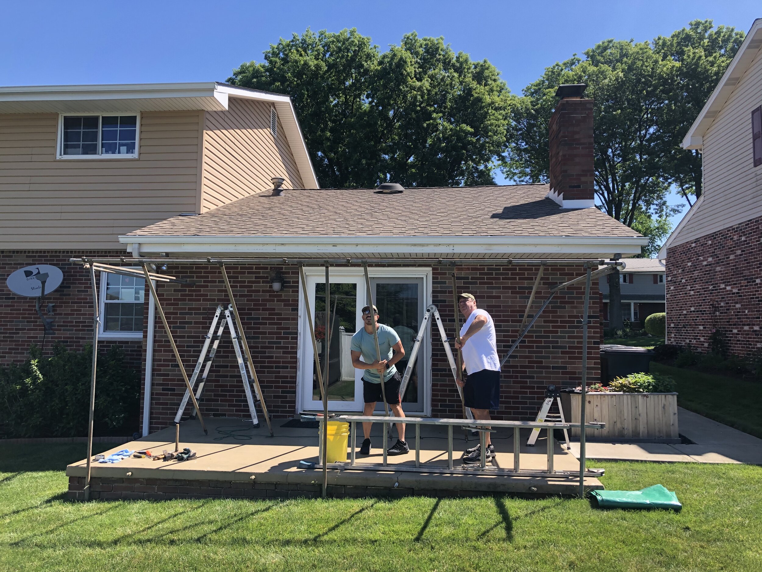Two men working on a wooden deck attached to a brick house, with ladders and tools around.