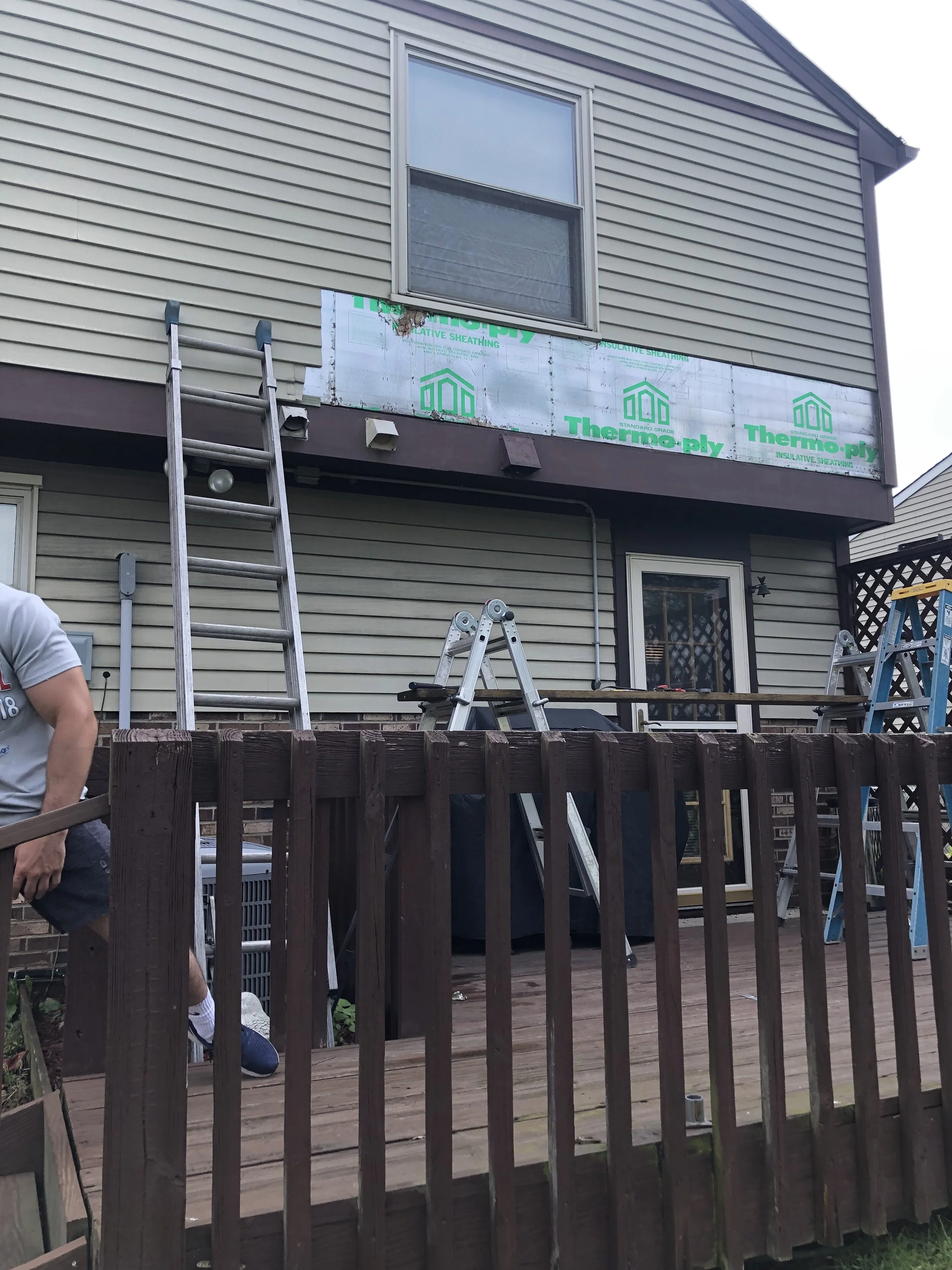 The image shows the exterior of a house with siding and a small screened door. There is a wooden deck with various ladders and tools set on it. A partially installed insulation and sheathing material is visible on the space between the upper and lowe