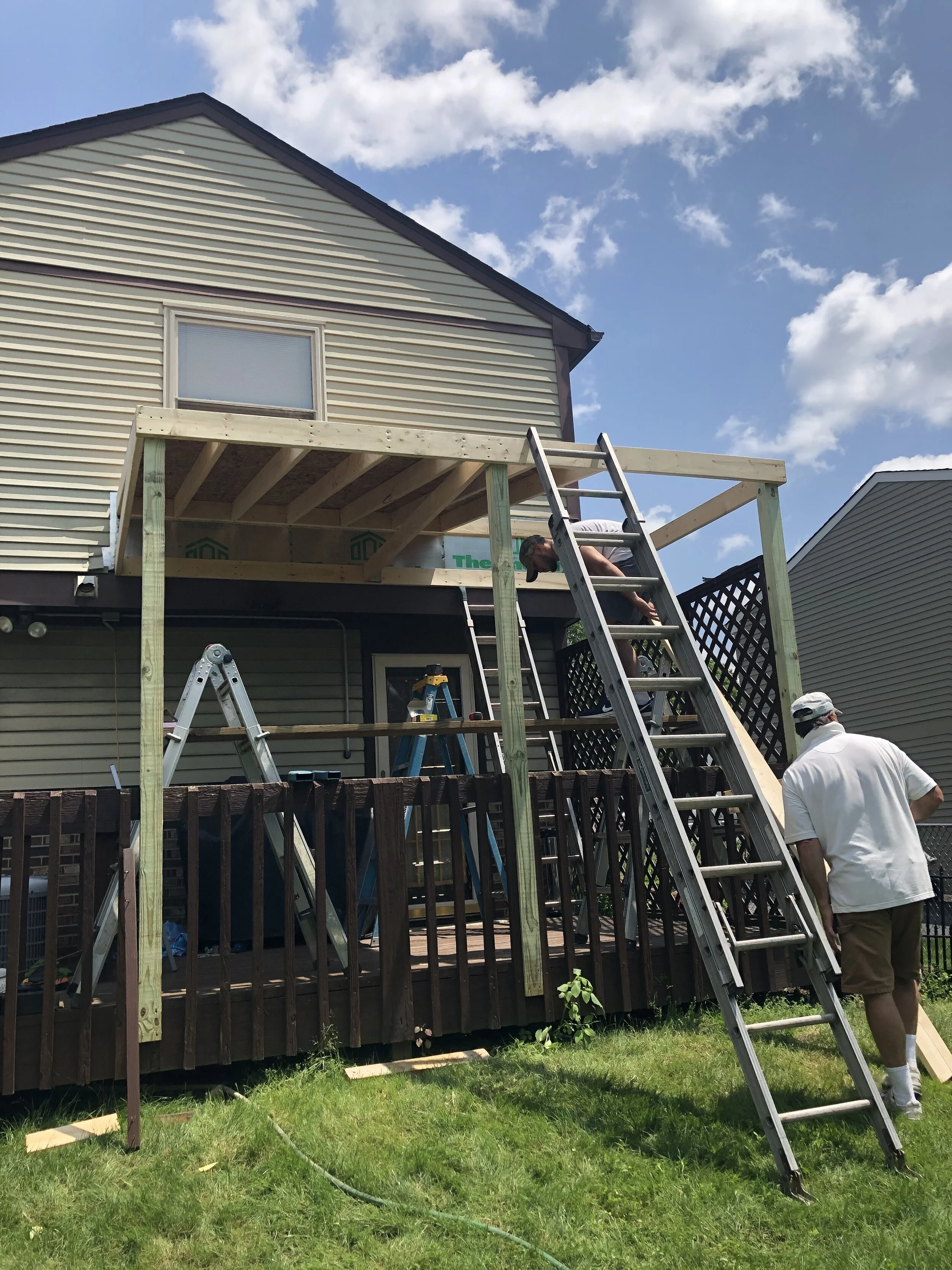 Two people are building a wooden deck extension at the back of a house, with ladders and construction tools around on a grass lawn.