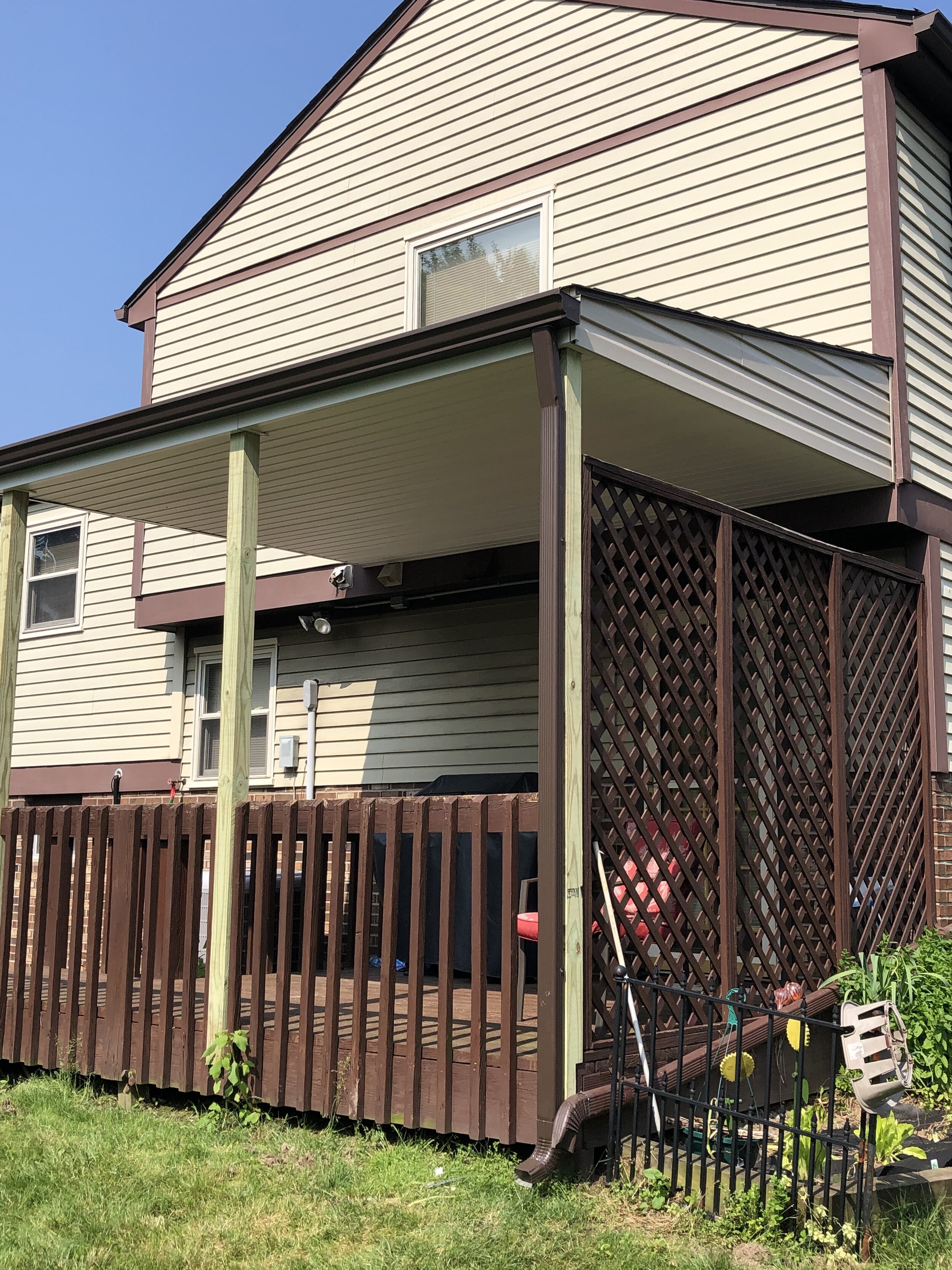 Backyard area with a newly built wooden deck and fenced enclosure. The house has beige siding with brown trim, and there is a lower covered patio area supported by green wooden posts. Gardening tools, a small bench, and a garden bed are visible.