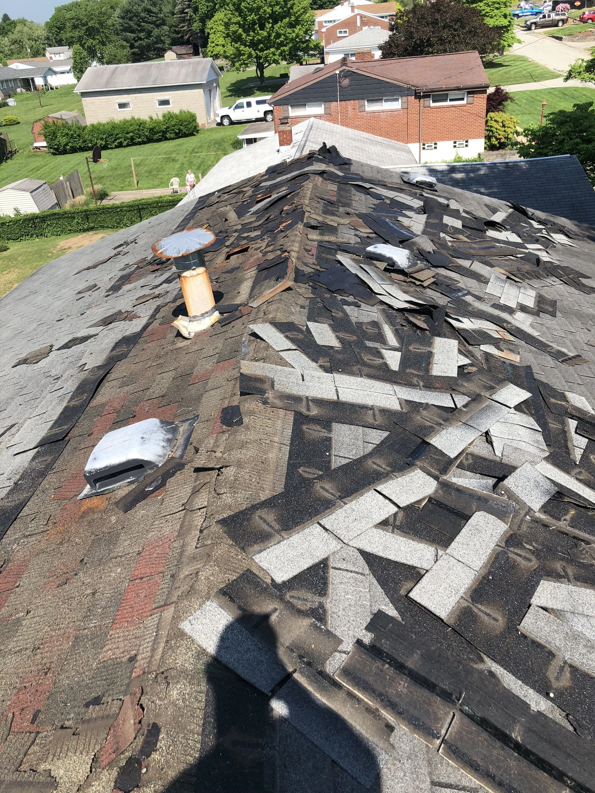 View of a damaged asphalt shingle roof with missing and displaced shingles, protruding roof vents, and surrounding residential houses with green lawns and trees.
