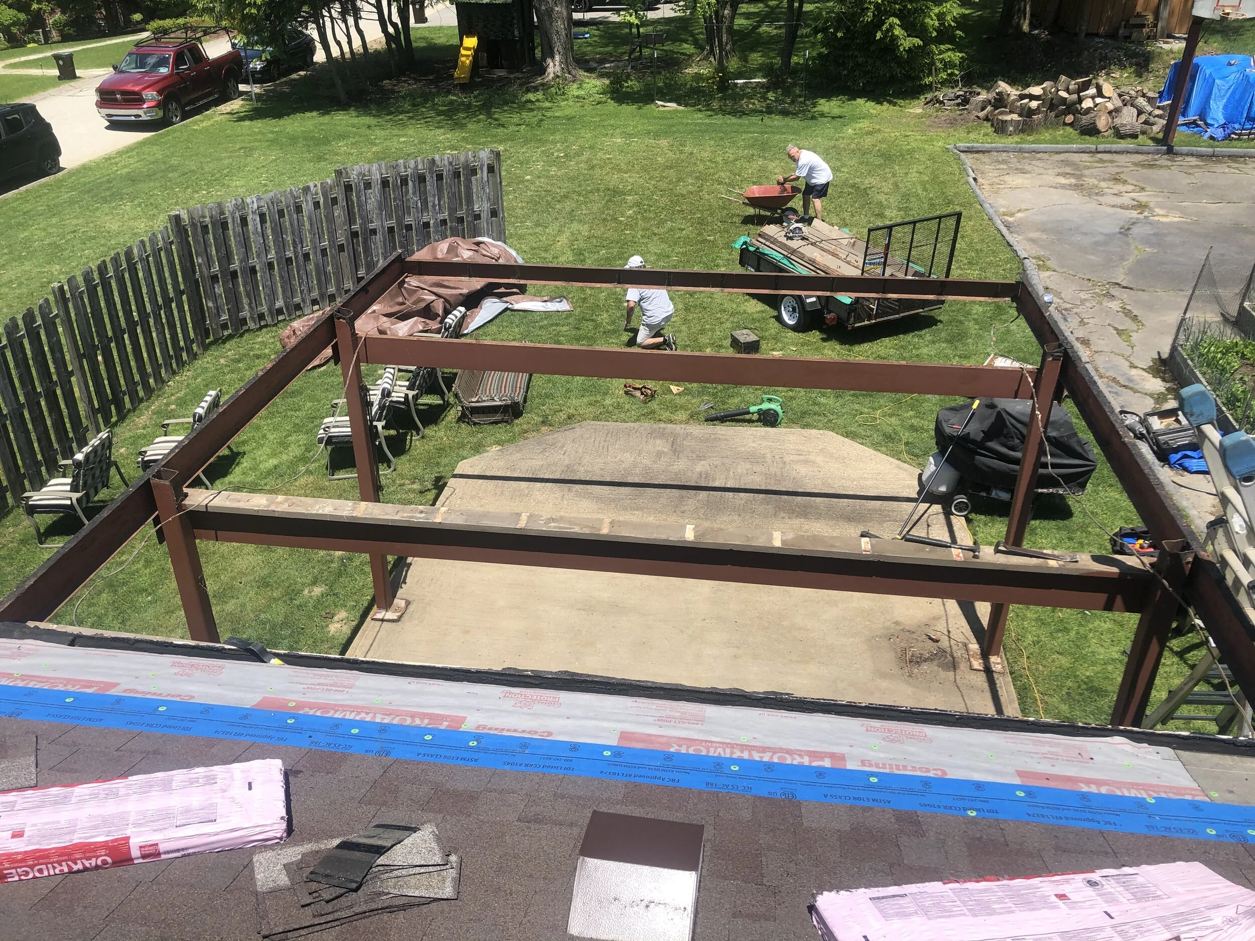 Two men working outdoors on a yard, one with a wheelbarrow and the other kneeling on the grass, with various tools and equipment around. There are cars parked and a wooden fence in the background.
