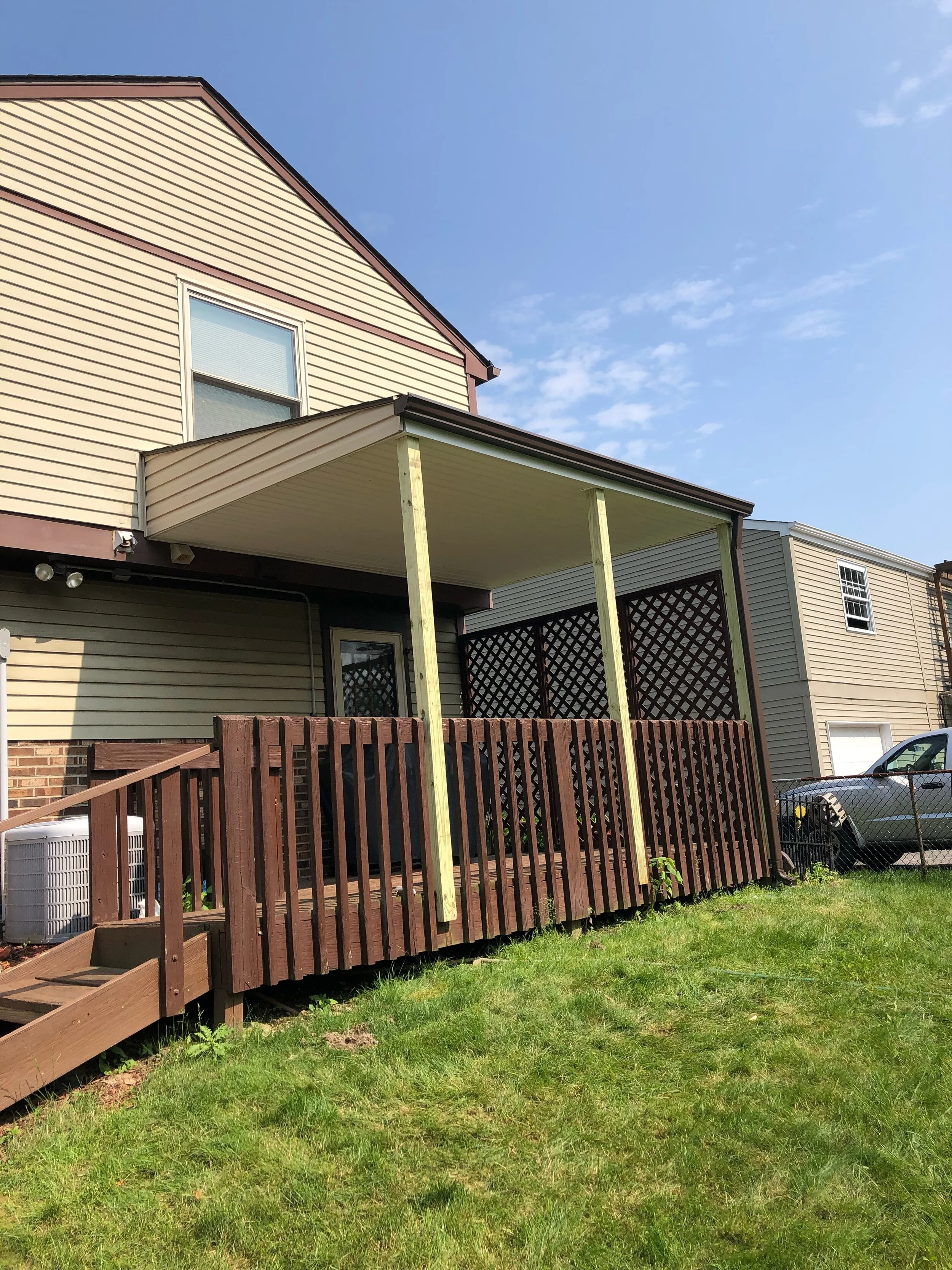 Backyard with a deck under construction, including wooden posts and railing, and a neighboring house with vinyl siding and a window, with a lawn and a car in the background.