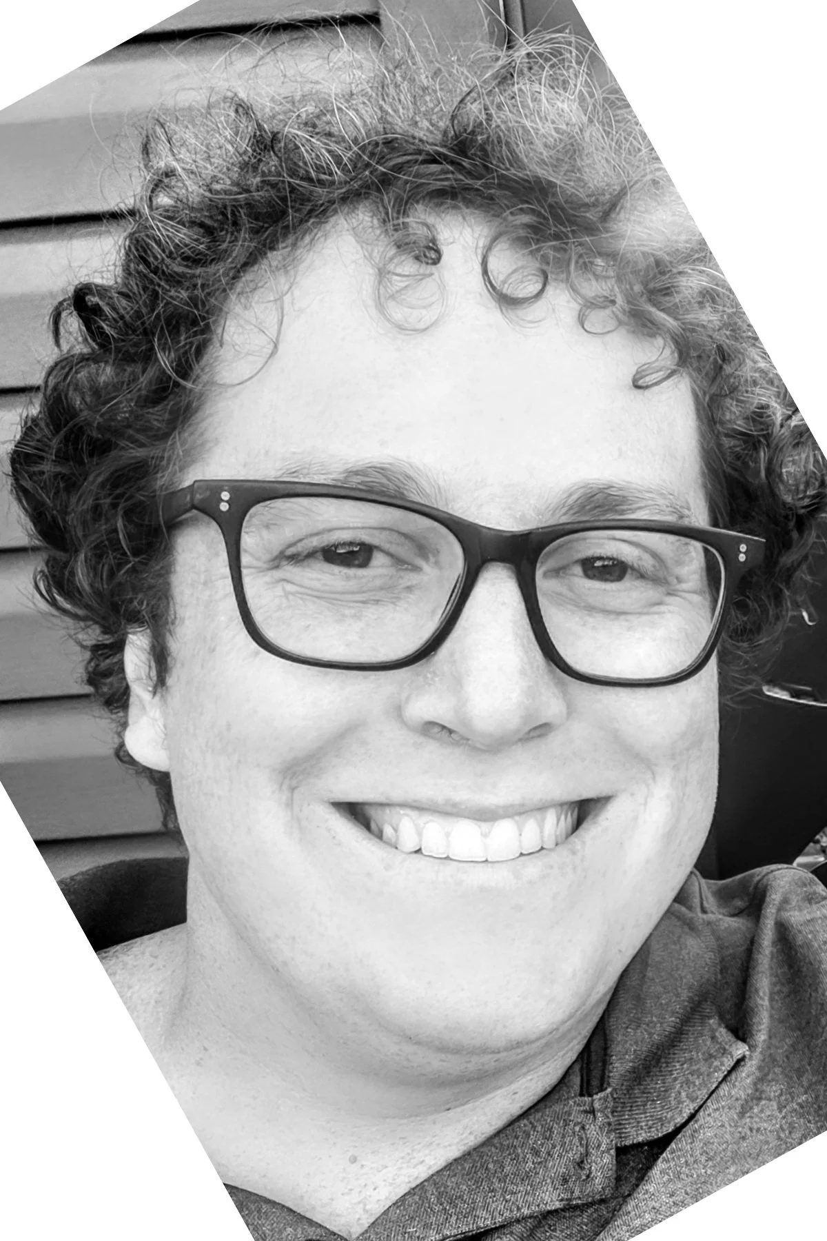 A person with curly hair and glasses smiling, wearing a hoodie, in black and white.