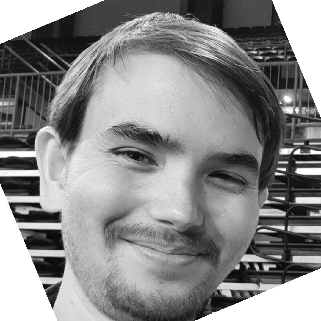 Close-up black-and-white photo of a young man with a slight smile, short hair, and a beard, in a venue with empty seats and railings in the background.