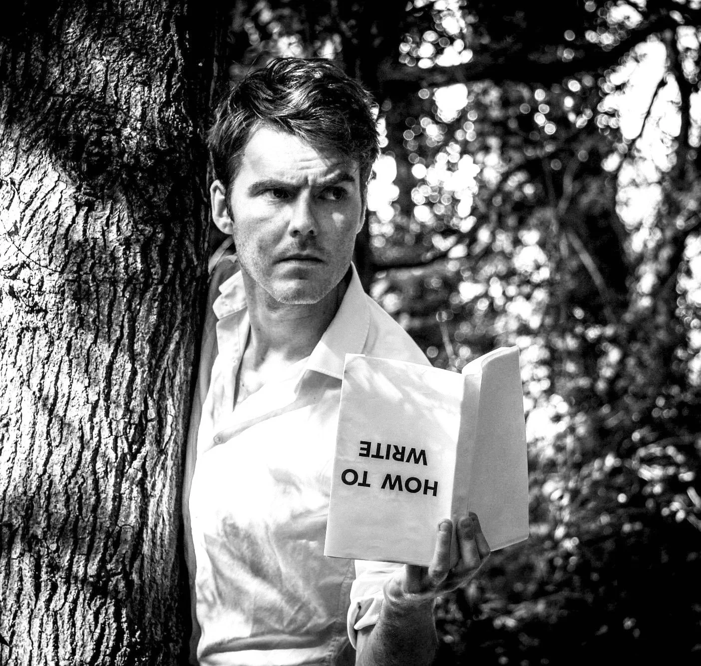 A black and white photo of a serious man leaning against a tree in a forest, holding a sign that reads 'HOW TO WRITE' in reference to the book.