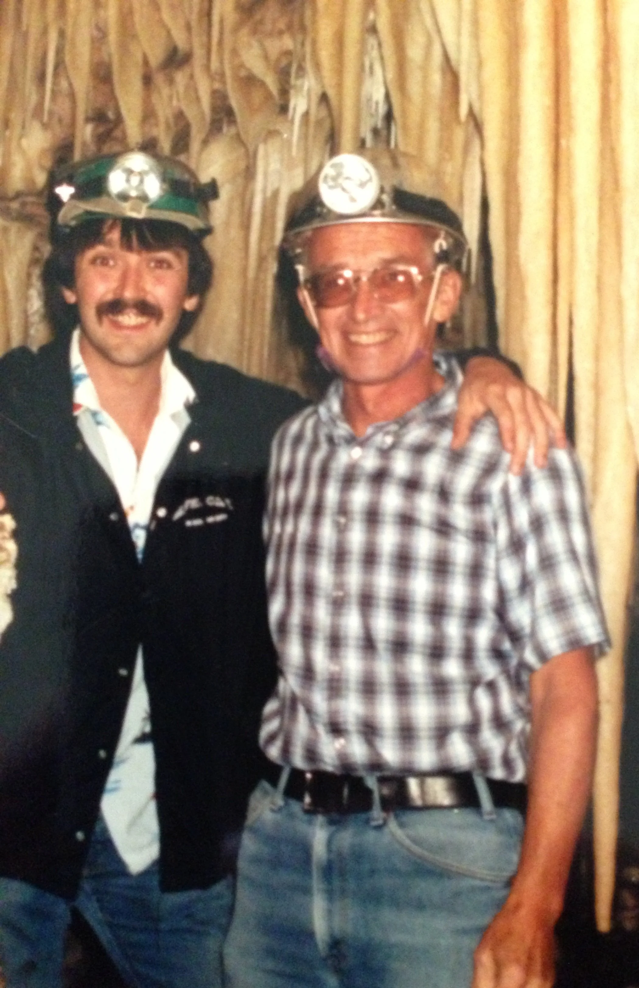 Two men wearing headlamps and casual clothing, standing together in front of a cave background, smiling.