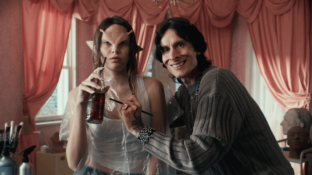 A person dressed as a fairy tale character with Elvish features holding a drink and a paintbrush, standing next to an older man with dark hair, smiling, in a room with pink curtains.