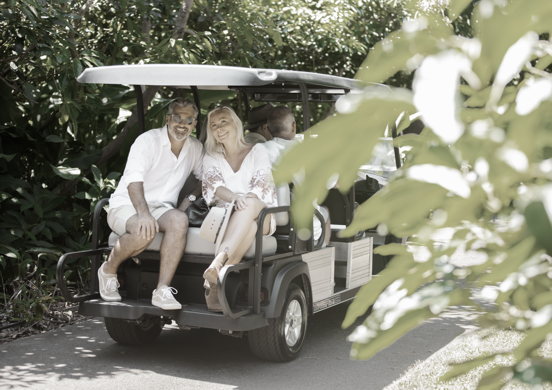 Four people ride in a golf cart on a shaded path surrounded by greenery, smiling and enjoying the outdoors.