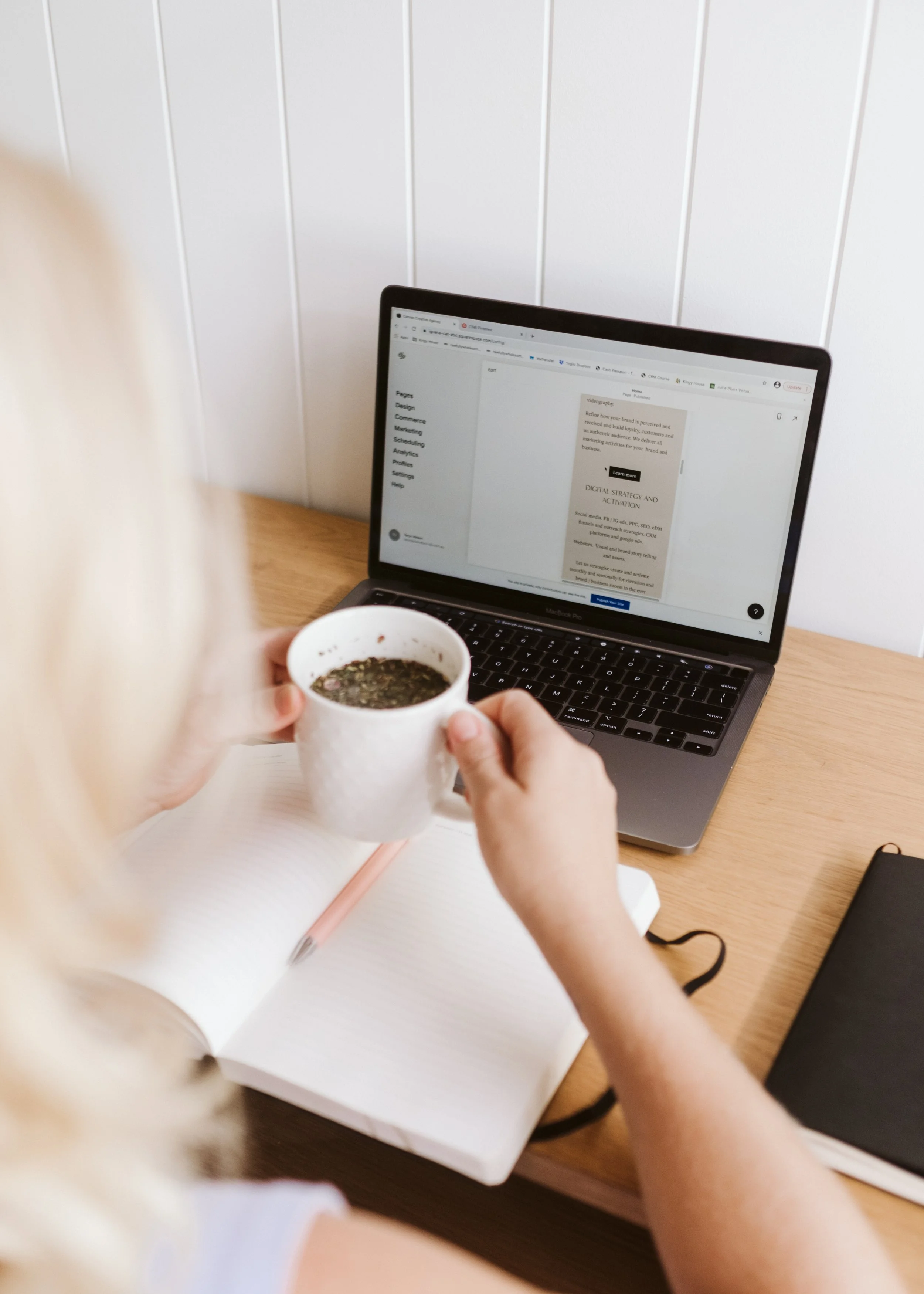 Person working on a laptop with a notebook and pen, holding a cup of tea or coffee, on a wooden desk with a black notebook nearby and a white wall in the background.