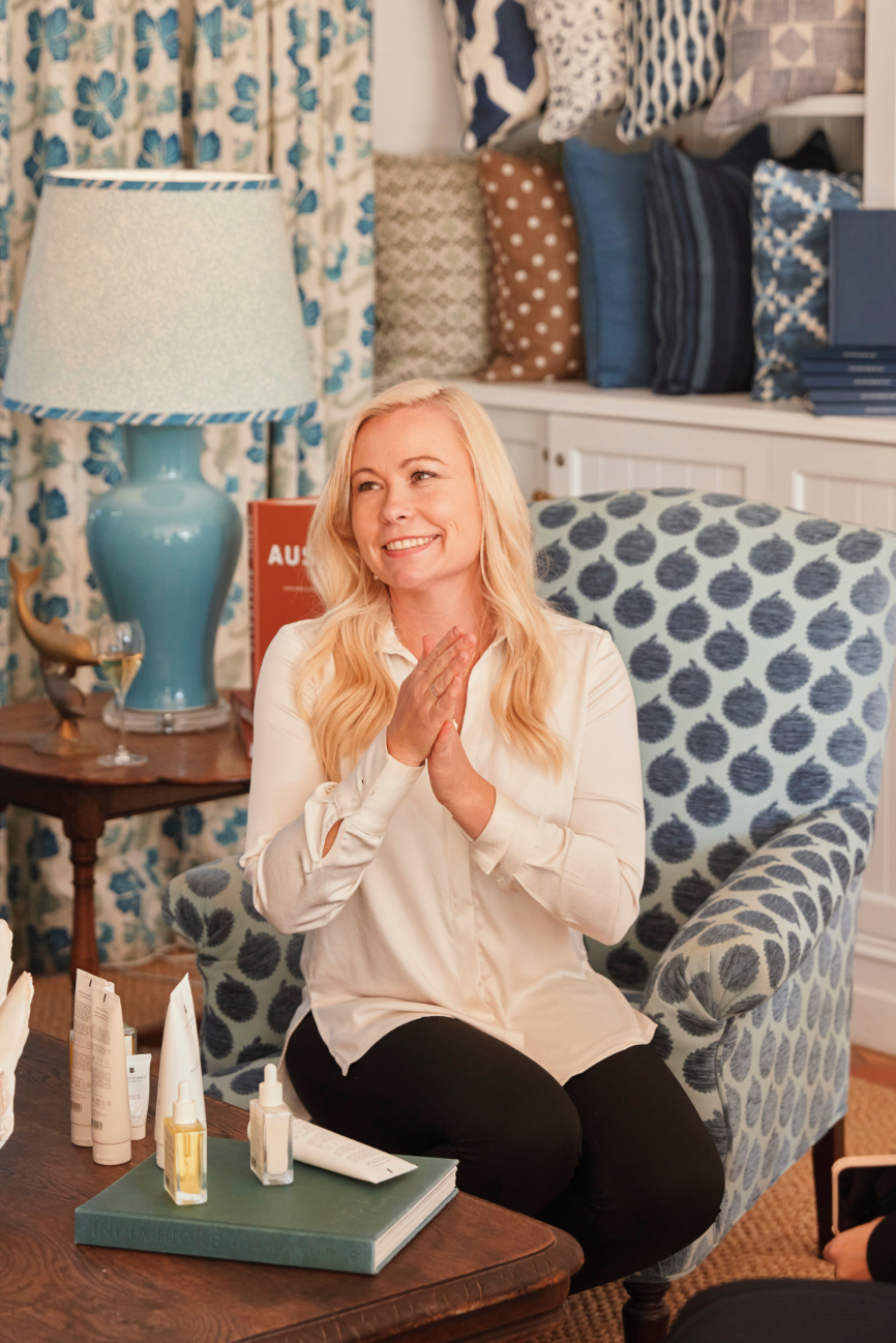 A woman with long blonde hair, wearing a white blouse and black pants, sitting in a cozy room with patterned cushions and books in the background. She is smiling and clasping her hands together.