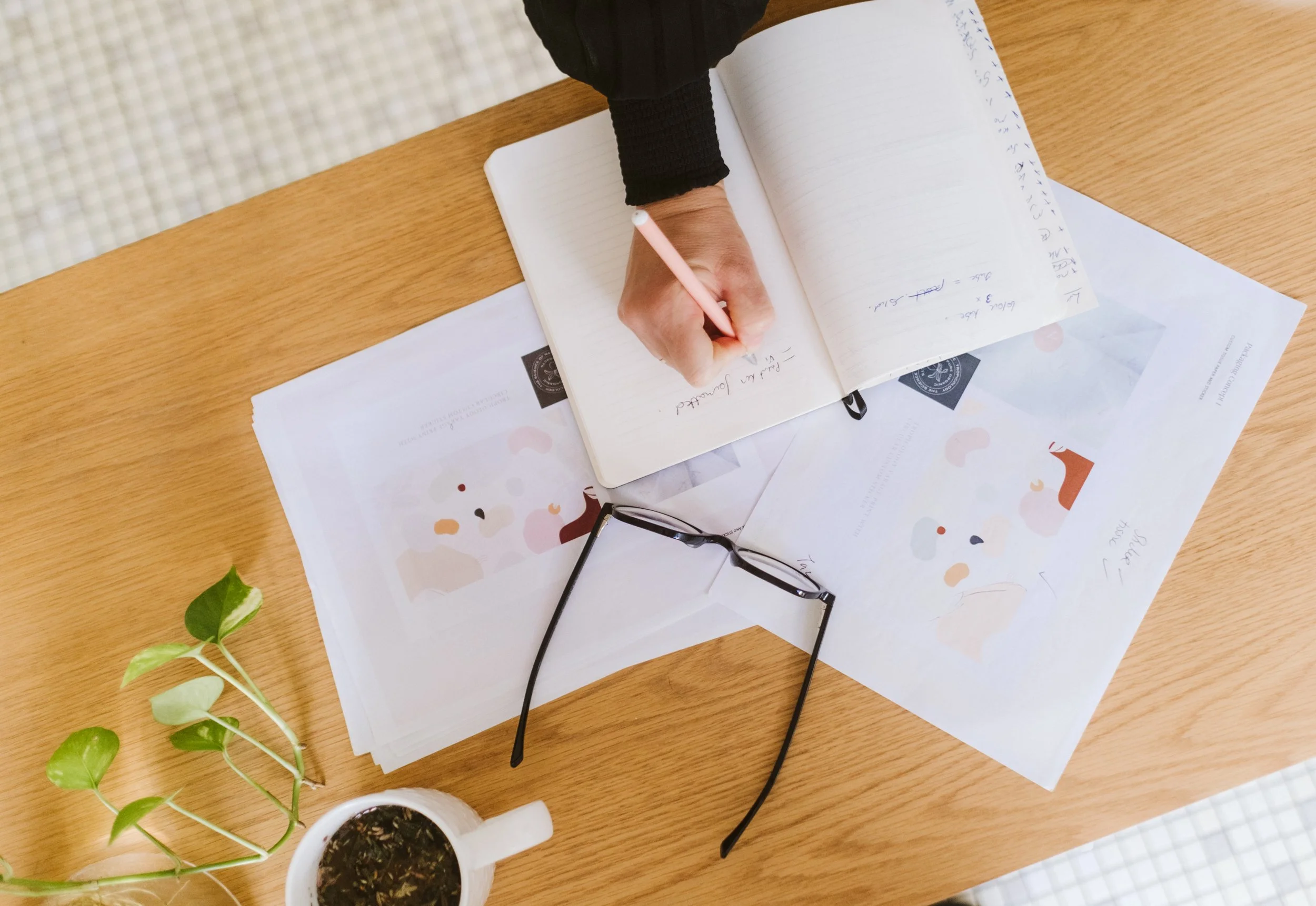 A person is writing in a notebook on a wooden table, with reading glasses, printed papers, a cup of tea, and a green plant nearby.