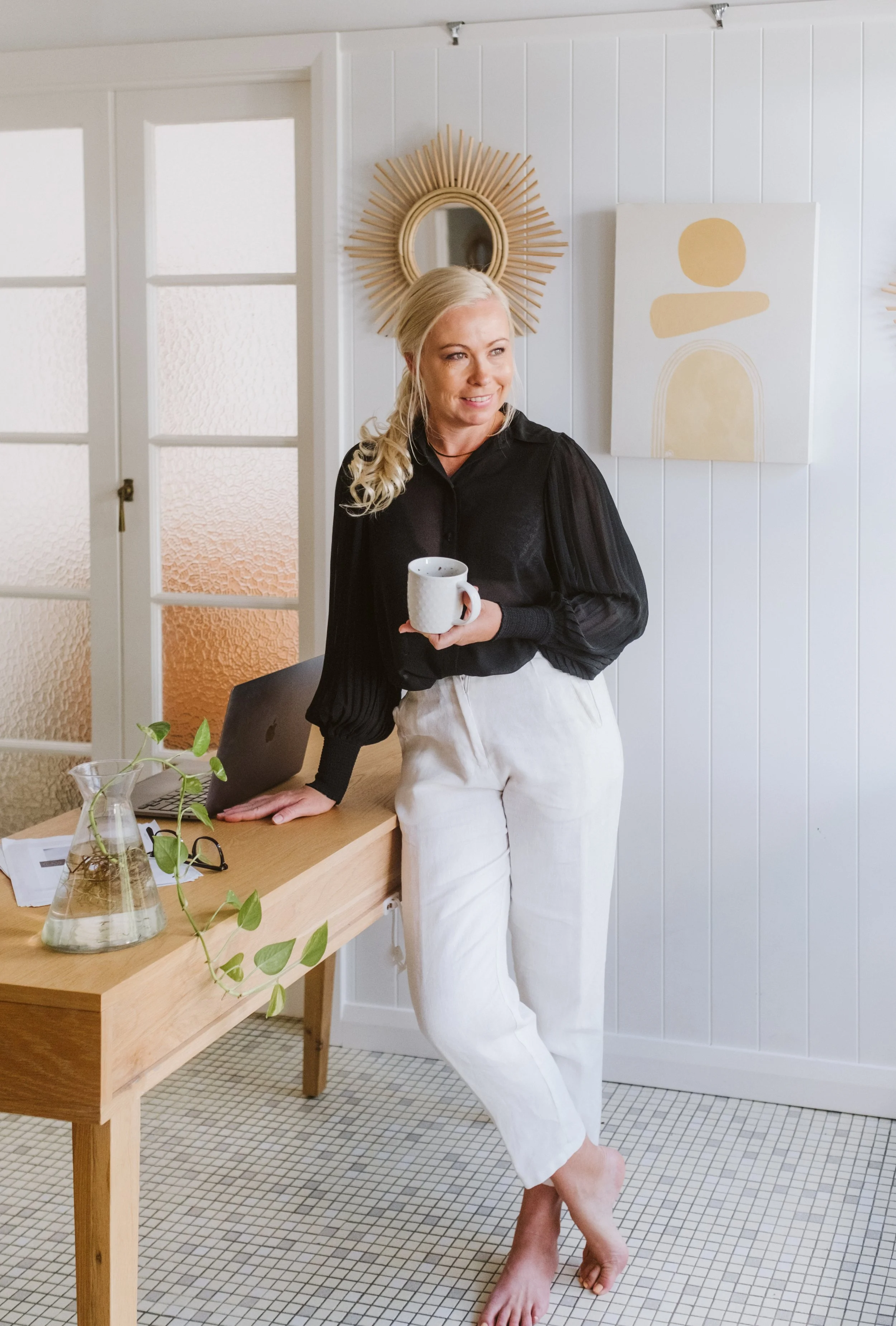Woman with blonde hair in a ponytail, wearing a black blouse and white pants, holding a mug. She is standing barefoot near a wooden table with a laptop, glasses, and a plant in a glass vase, in a bright, modern room with white paneled walls and decorative artwork.