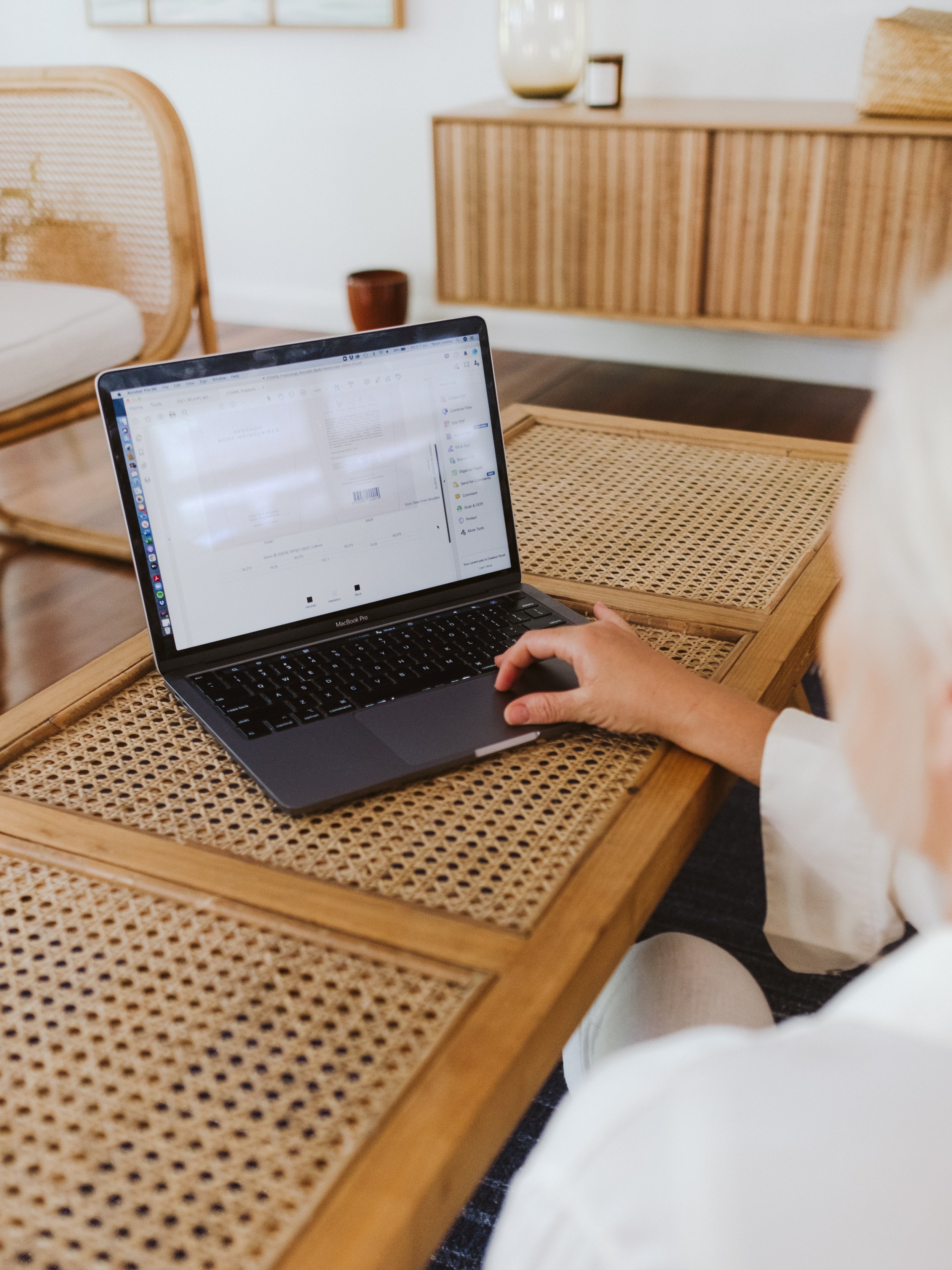 Person using a MacBook Pro laptop on a wooden woven table in a modern, minimalist living room with wooden furniture and decor.
