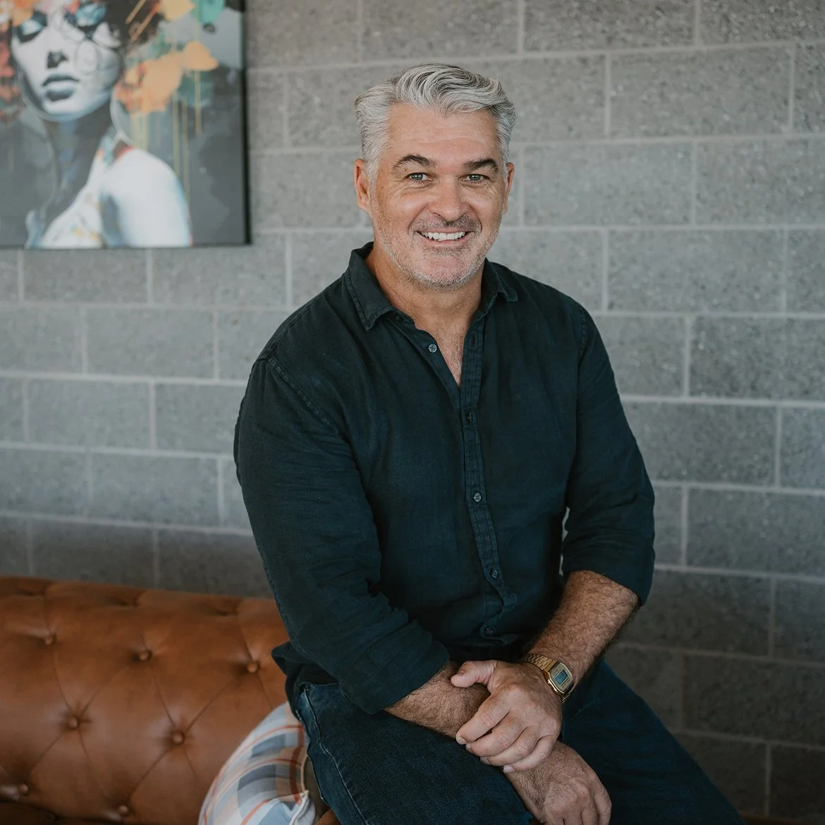 A smiling middle-aged man with gray hair and a beard, wearing a black shirt, sitting on a brown tufted leather bench in front of a gray brick wall with artwork.