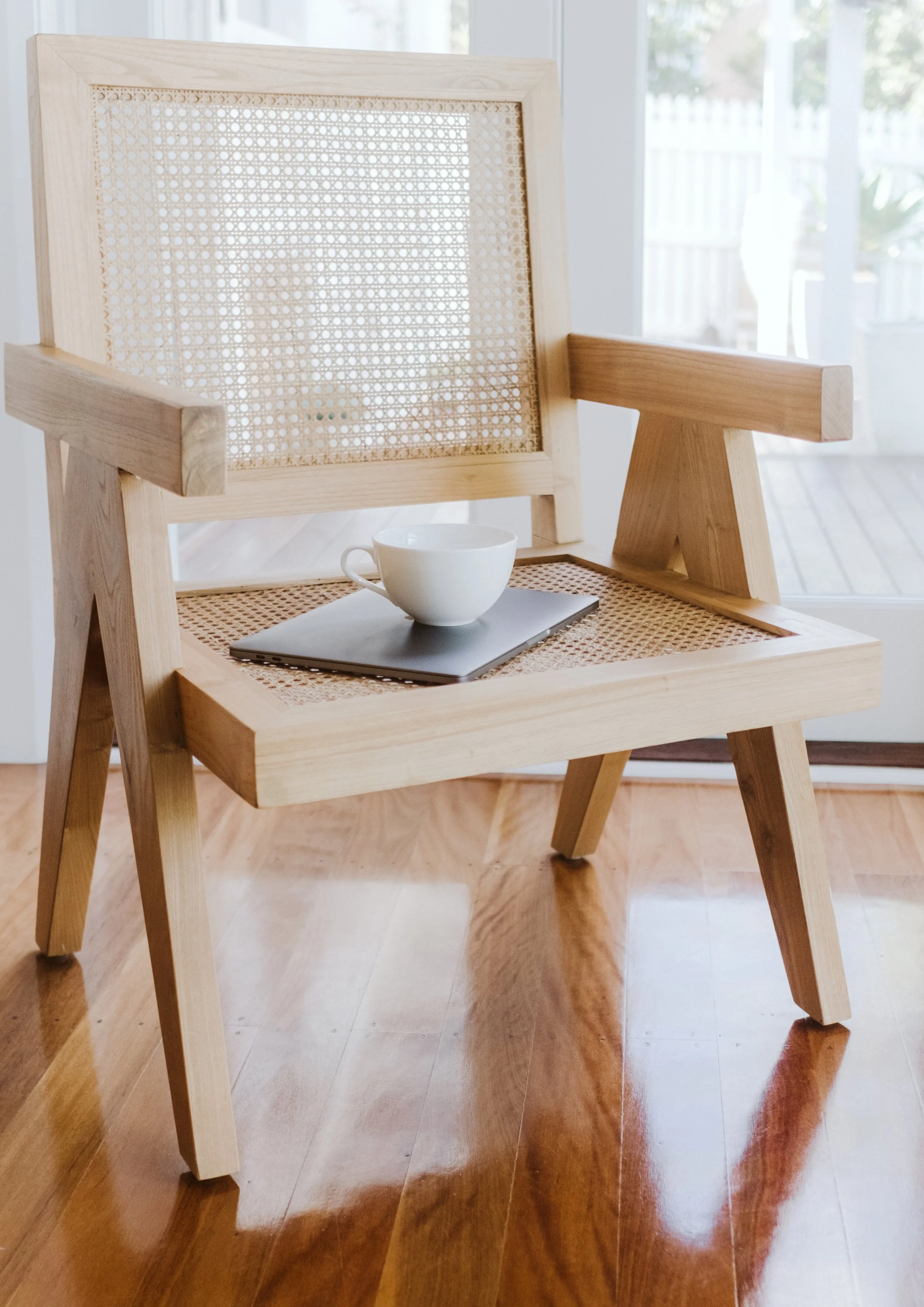 A wooden chair with a cane seat and backrest, placed indoors with a glass door in the background. On the seat, there is a closed laptop and a white coffee cup.