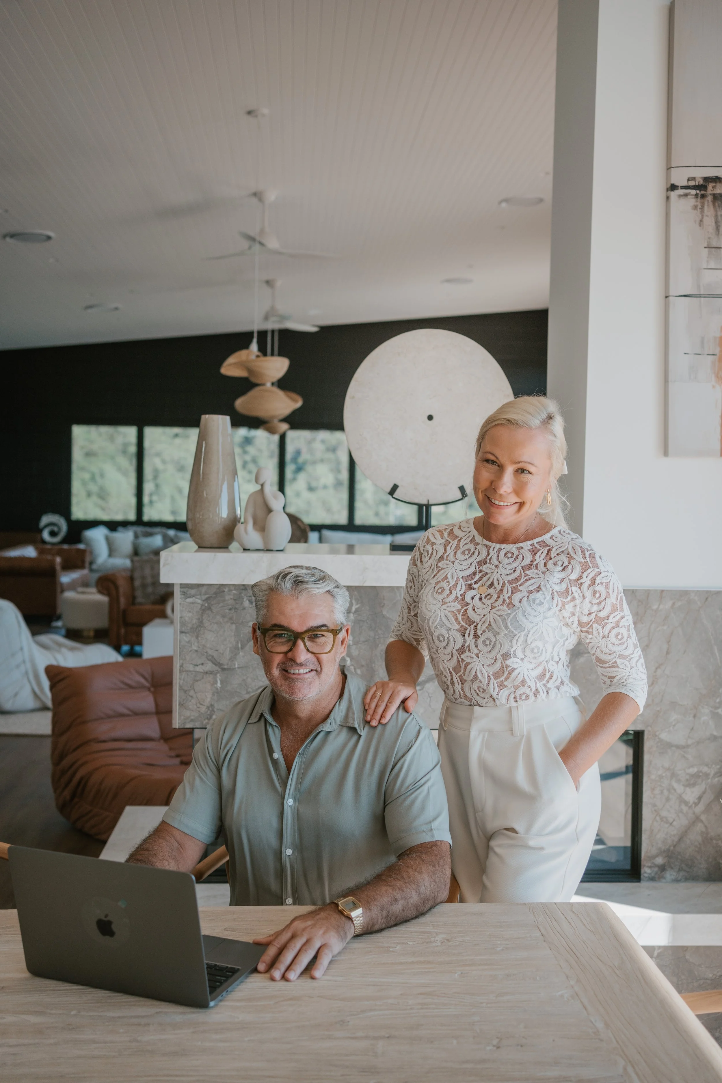 A smiling woman with blonde hair, wearing a lace white top, standing next to a seated man with gray hair and glasses, working on a laptop at a table in a modern, well-lit living room.