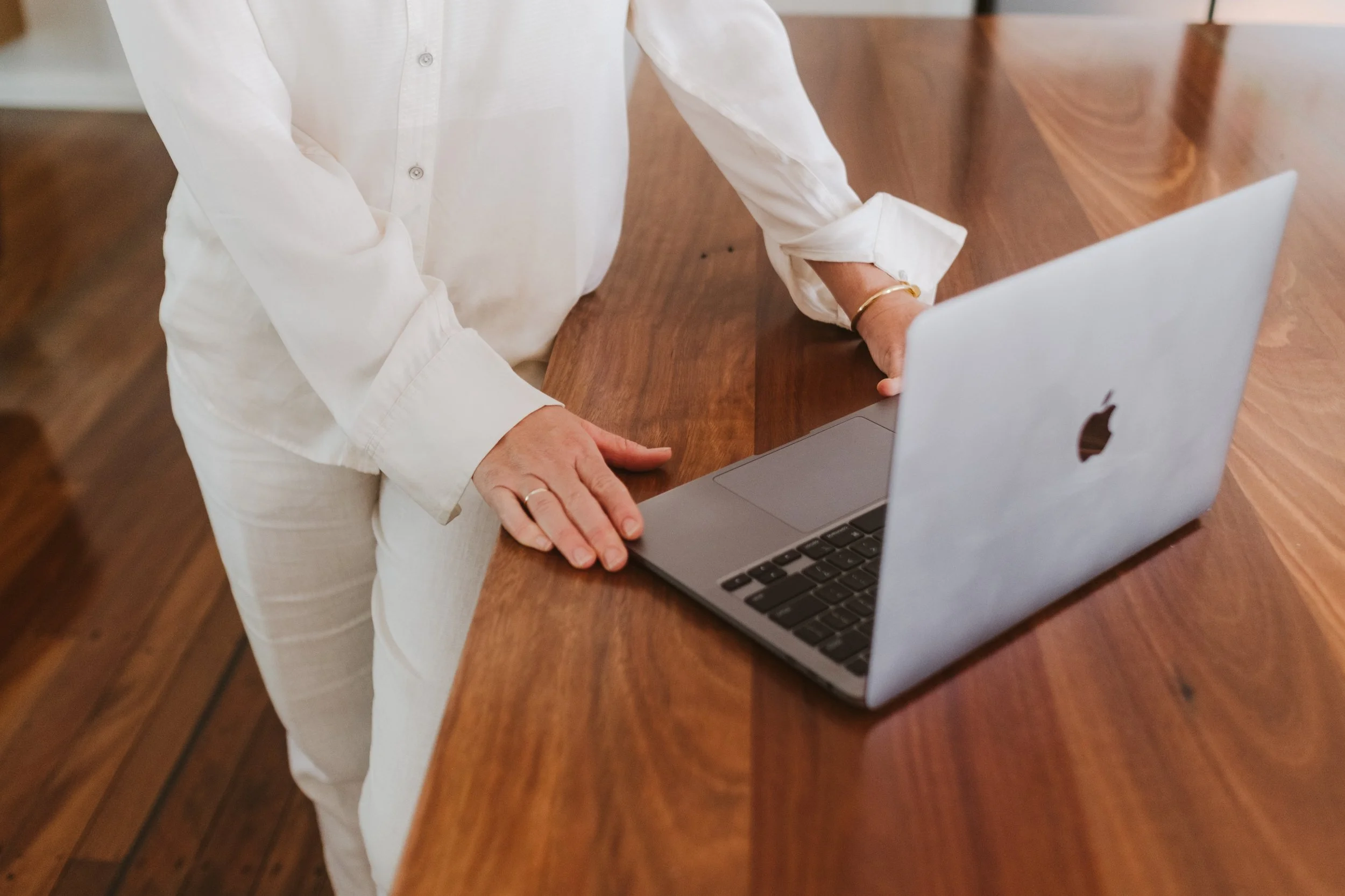Person in white clothes leaning on a wooden table with hands resting on a silver MacBook laptop.