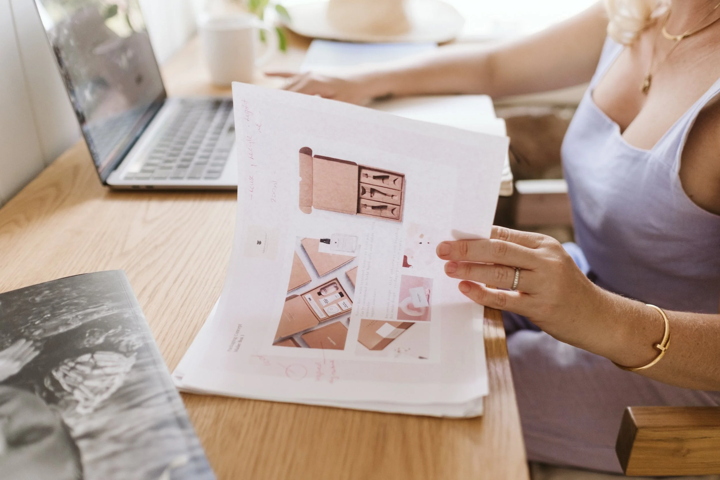 Woman sitting at a wooden table reading printed pages with interior design images, laptop, and a cup in the background.